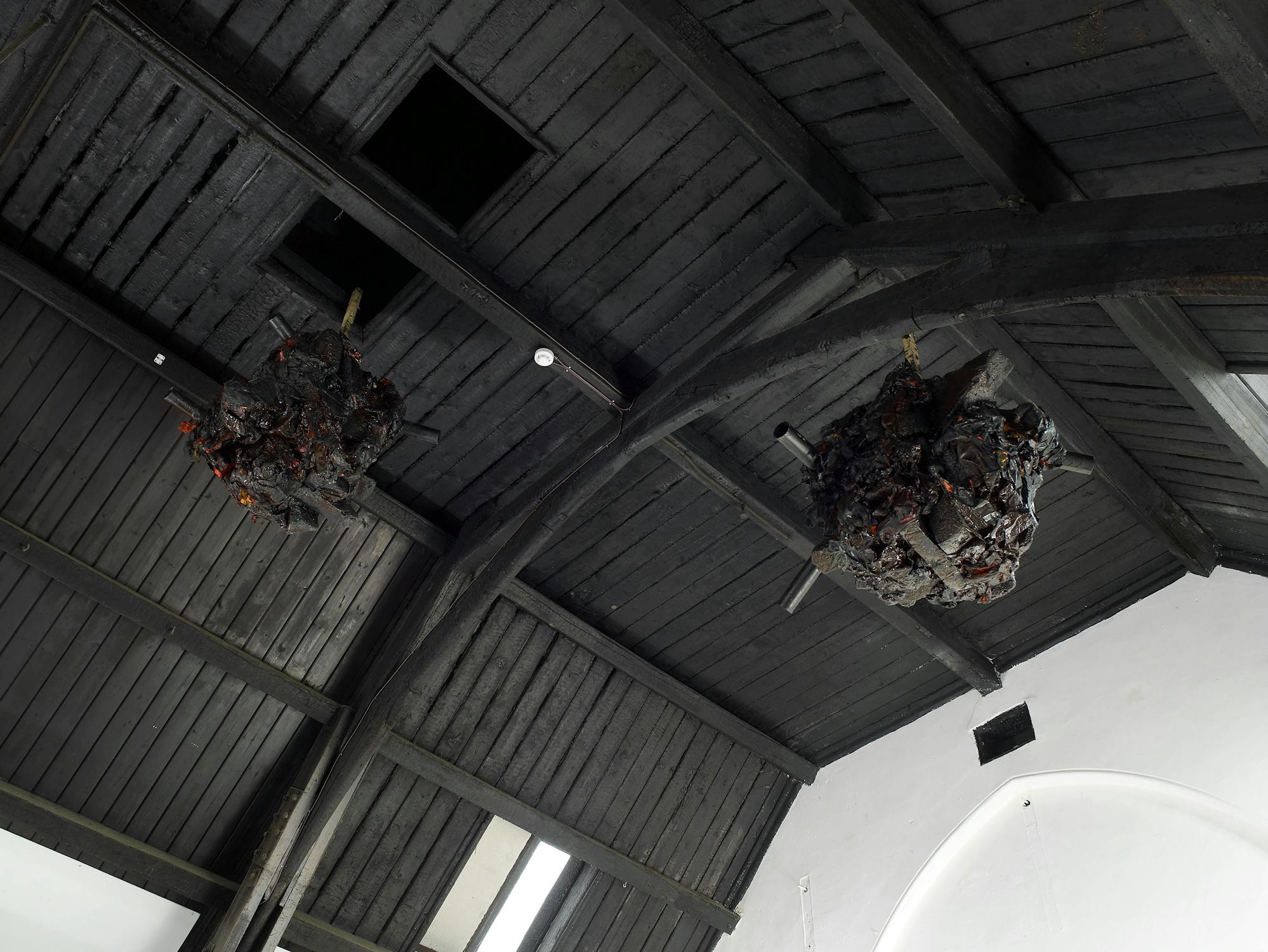 A view looking up at a dark wooden ceiling with exposed beams. Two large, irregularly shaped, dark sculptures hang from the ceiling, appearing charred or burnt, with some orange highlights resembling embers or molten material.