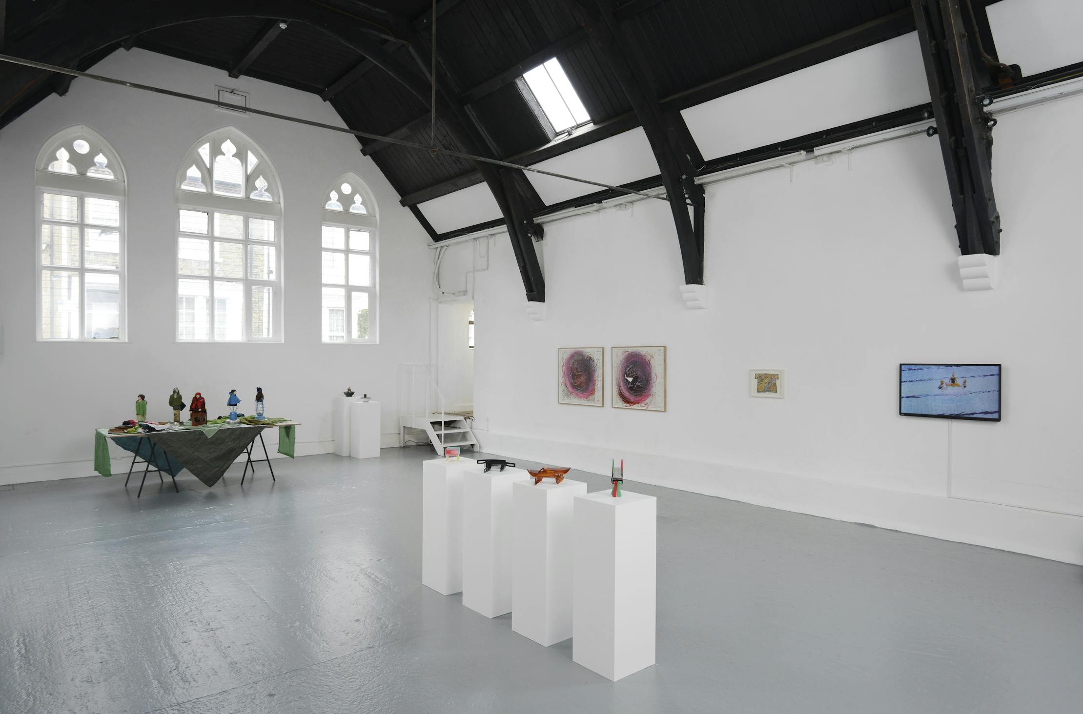 A bright, modern art gallery with white walls, tall arched windows, and a dark wooden ceiling. Artworks, sculptures on pedestals, and a table with colourful objects are displayed throughout the space.