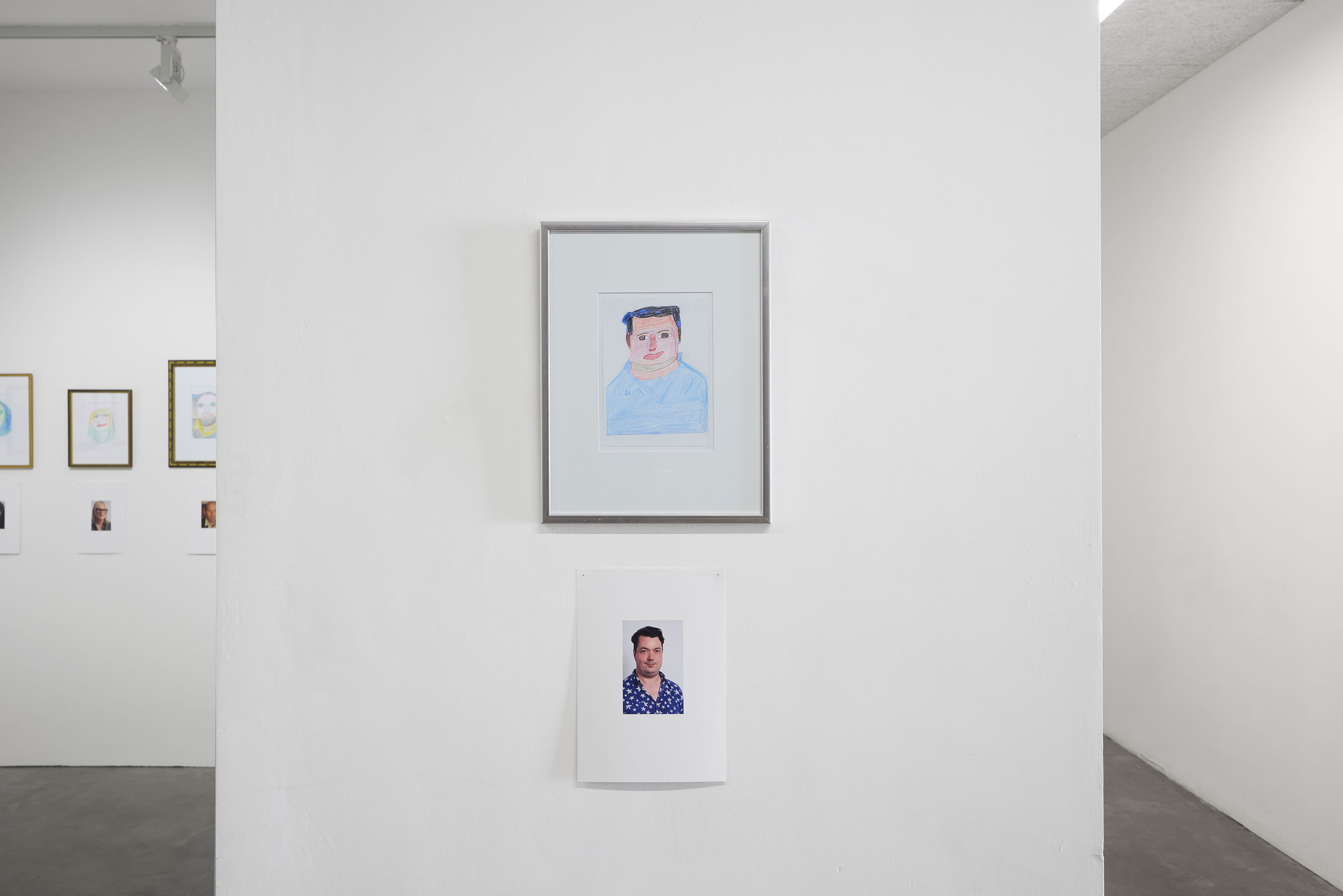 A framed drawing of a man (Joe Scotland) hangs above a photo of the same man wearing a blue patterned shirt; both are displayed on a white wall in a minimalist gallery, with additional framed art partially visible on adjacent walls.
