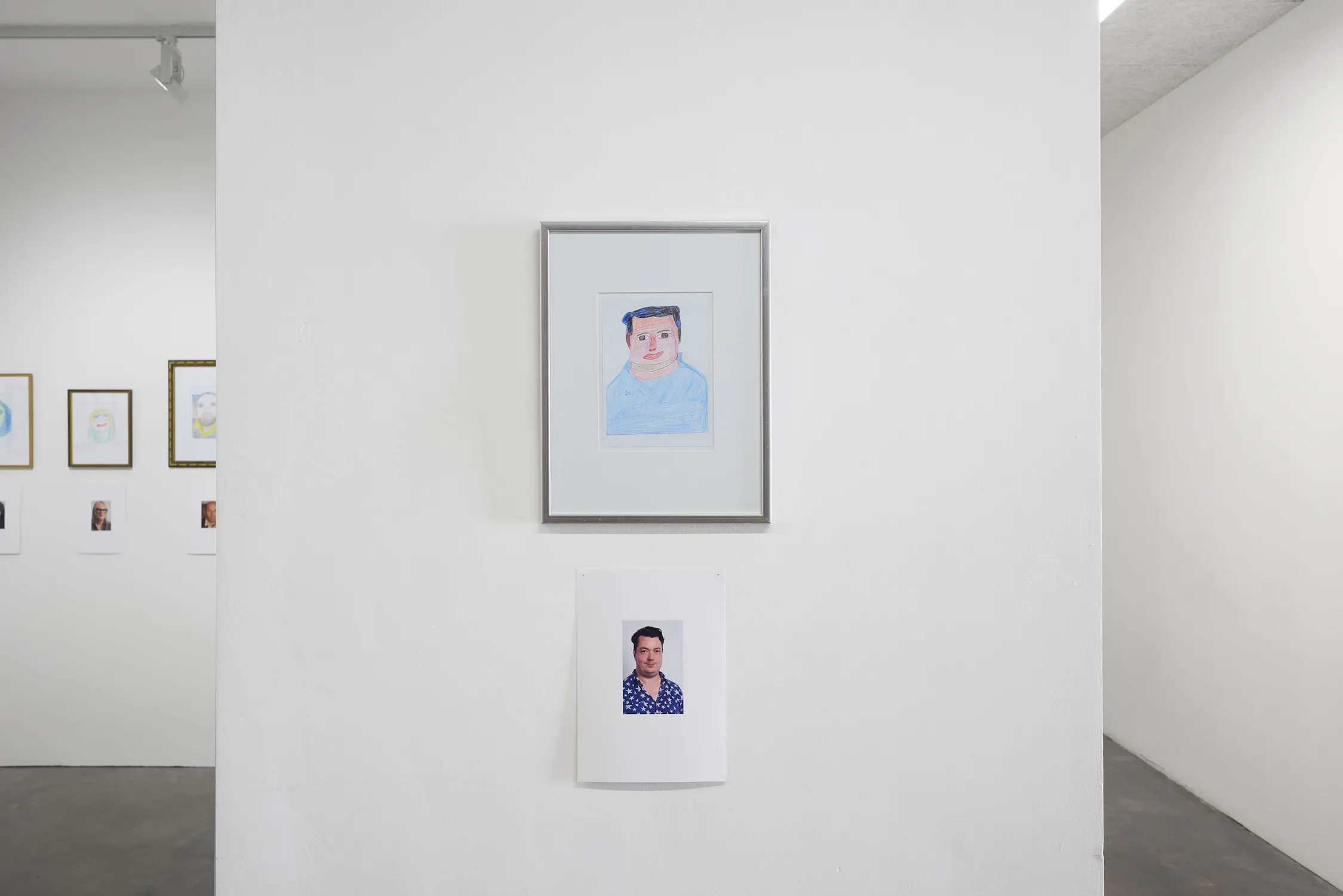 A framed drawing of a man (Joe Scotland) hangs above a photo of the same man wearing a blue patterned shirt; both are displayed on a white wall in a minimalist gallery, with additional framed art partially visible on adjacent walls.