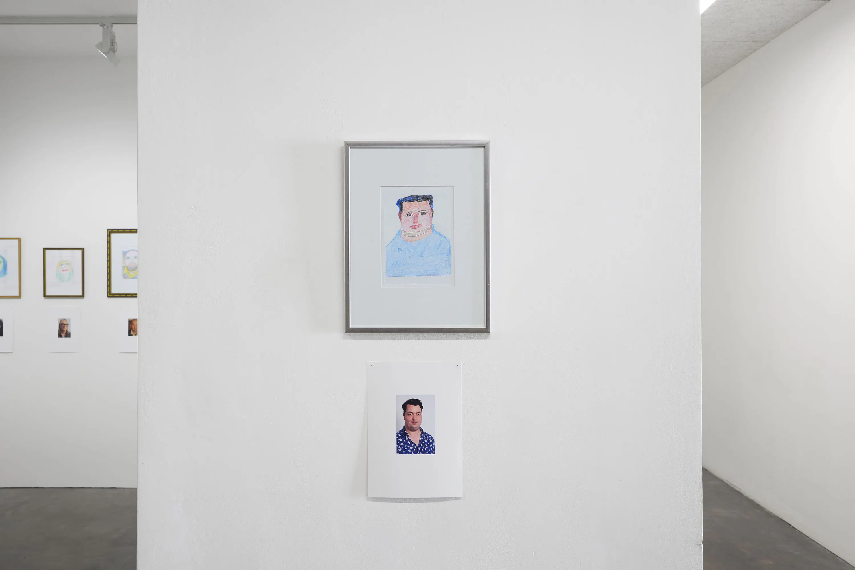 A framed drawing of a man (Joe Scotland) hangs above a photo of the same man wearing a blue patterned shirt; both are displayed on a white wall in a minimalist gallery, with additional framed art partially visible on adjacent walls.