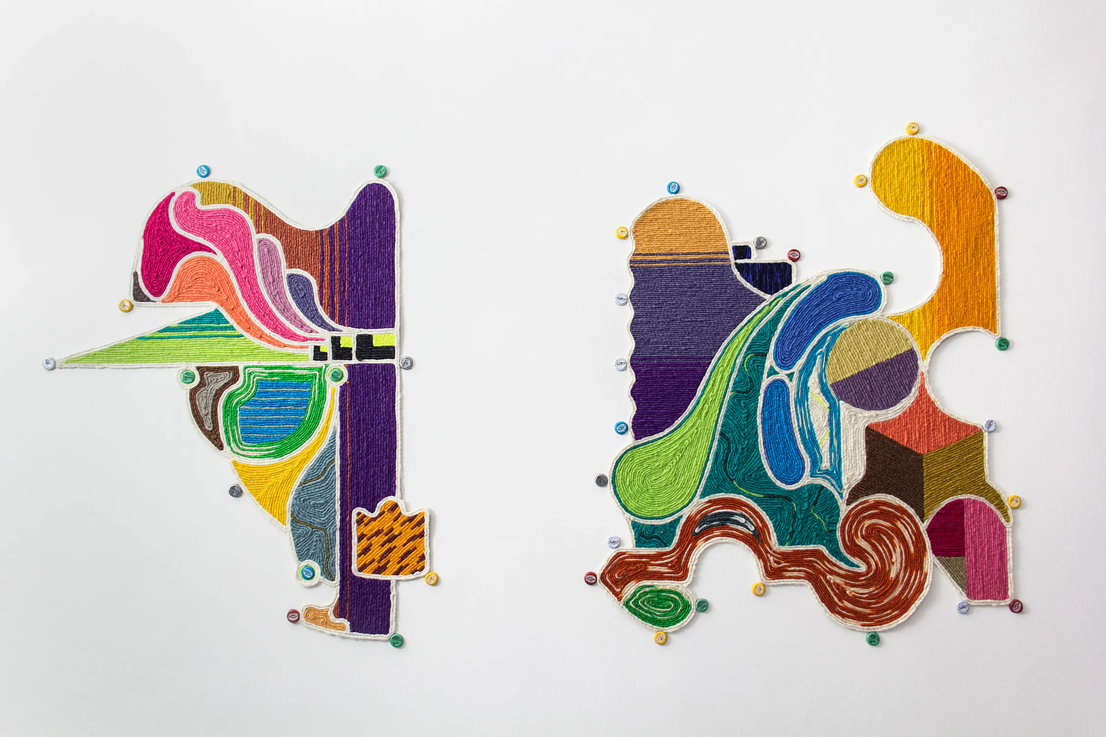 Two abstract, colourful embroidered woven textiles by Guido Yannitto are pinned to a white background. The left shape features bold curves and lines in pink, purple, green, and yellow. The right shape is larger, with swirling patterns in teal, blue, brown, gold, and red tones.