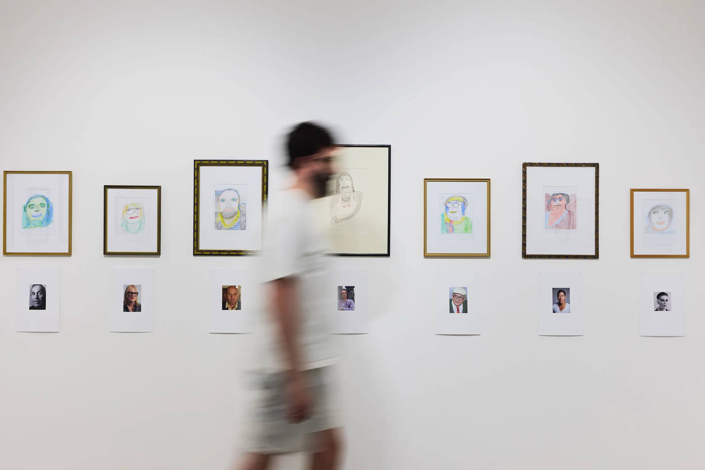 A blurred person walks past a gallery wall displaying framed colour pencil portraits. Beneath each drawing is a printed photo of a real person. The background is a plain white wall, creating a minimalist exhibition setting.