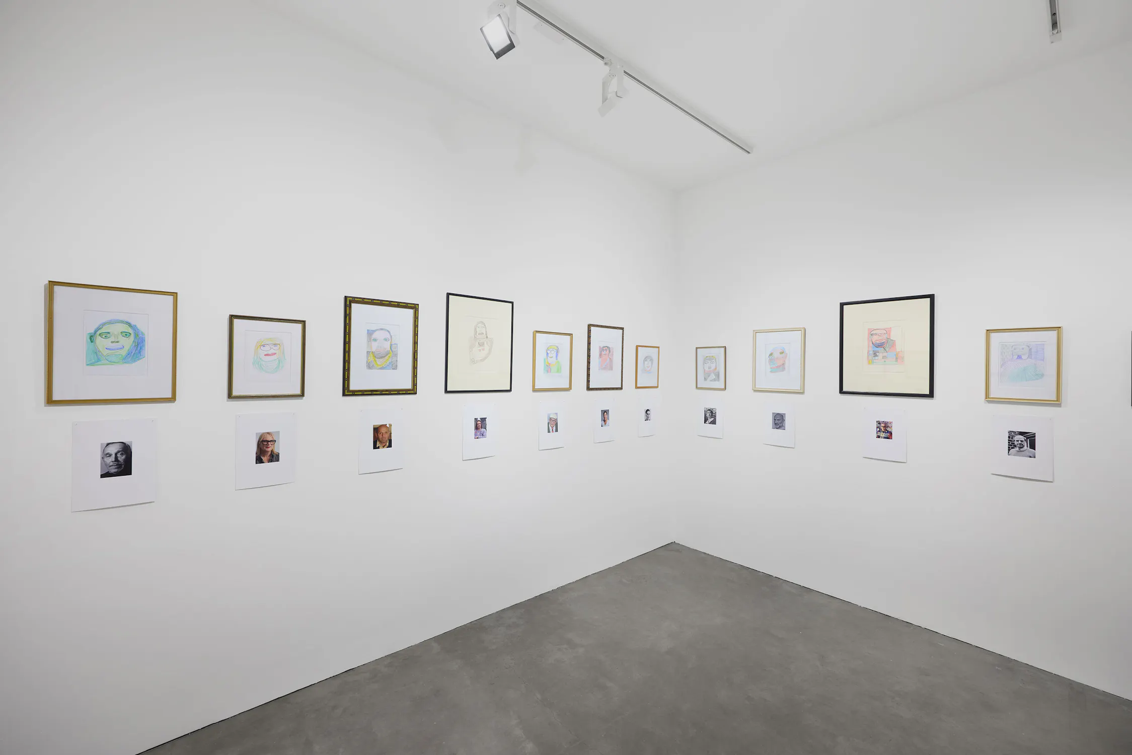 A white-walled gallery corner displays framed drawings of faces and lined-up printed photos below. The drawings and photos are evenly spaced in two rows on both walls, with minimal decor and a grey concrete floor. Ceiling lights illuminate the artwork.