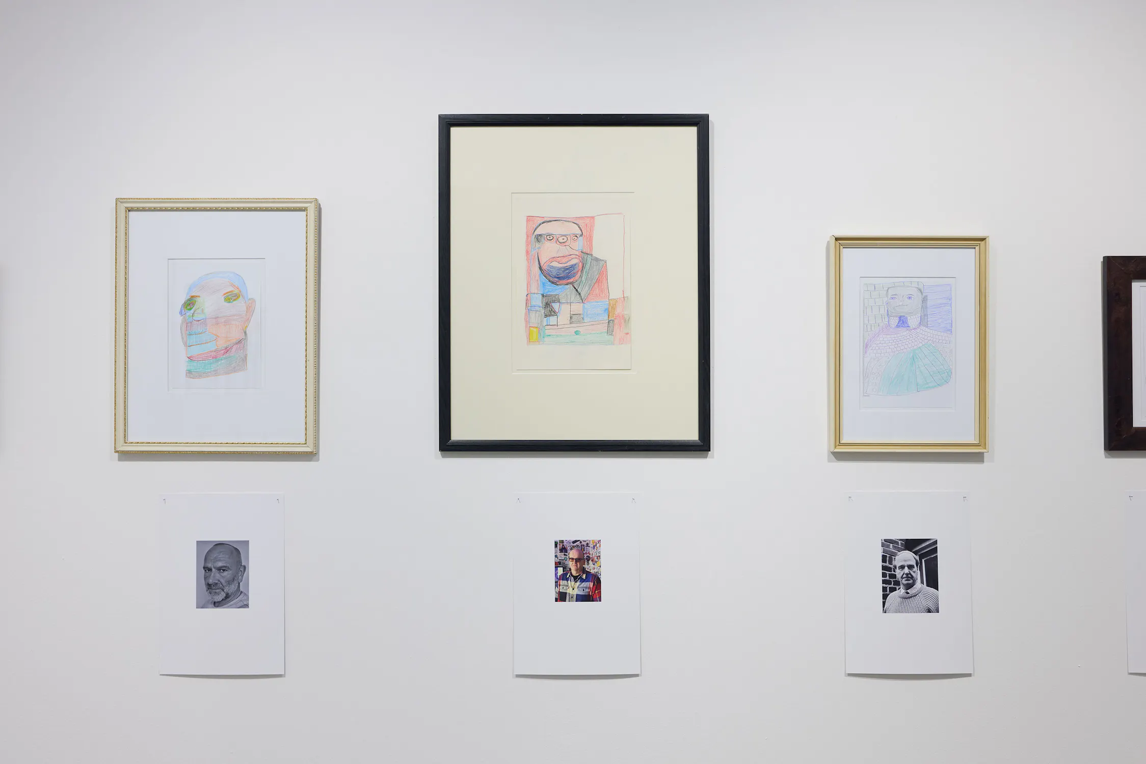 Three colourful portraits in frames hang on a white wall, each above a reference photo. The centre frame is larger and bordered in black, while the left and right frames are gold and silver, respectively. The portraits are hand-drawn with coloured pencil.