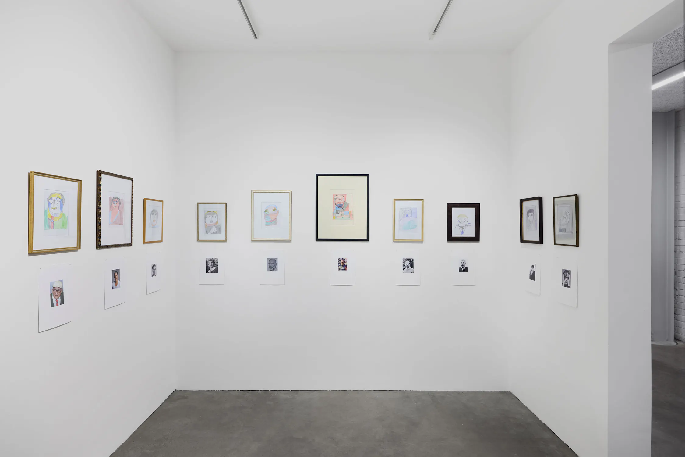 A white gallery corner displays framed portraits on two walls. Under each are small colour photos on paper. The floors are concrete and the lighting is bright and minimal, emphasising the art. The scene is clean, modern, and orderly.