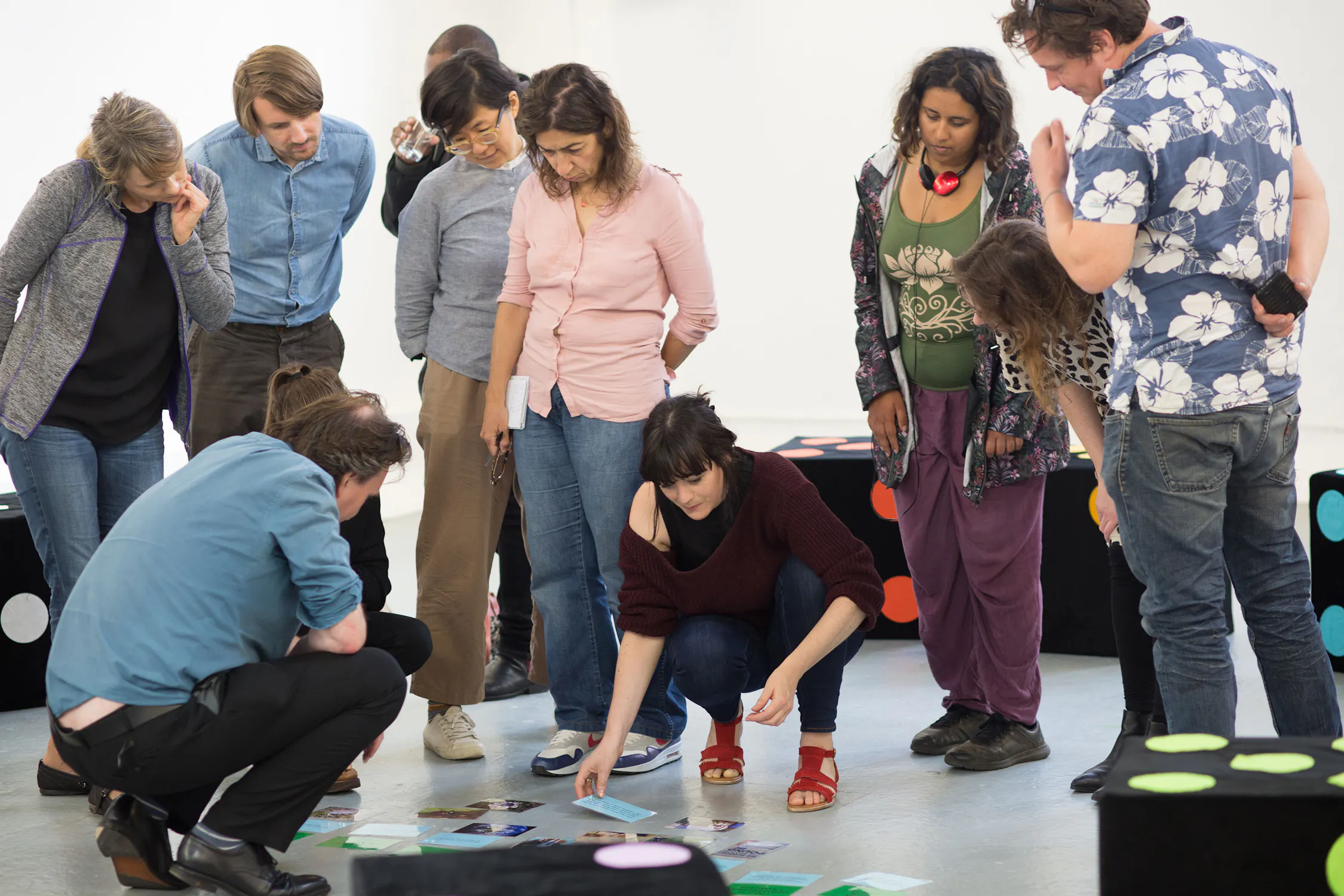 A diverse group of people stands in a circle watching as a woman kneels and arranges cards or images on the floor. Some participants lean in and observe closely. The room is bright, with benches featuring large coloured dots.