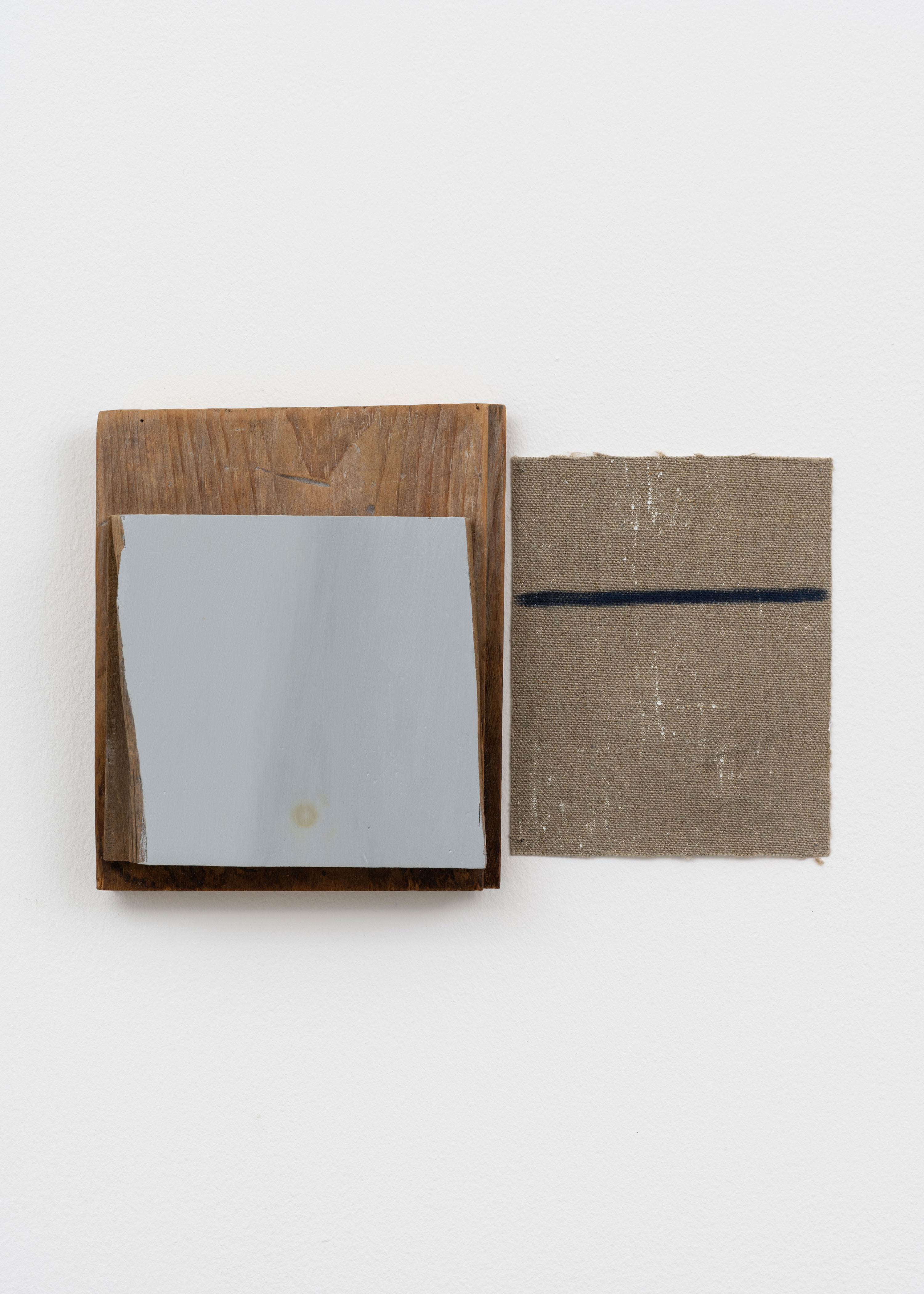 An abstract wall sculpture consists of a square of grey-painted wood layered over a dark wooden plank, positioned next to a square of raw, textured tan canvas featuring a single thin horizontal navy blue line. The pieces are mounted on a plain white wall.