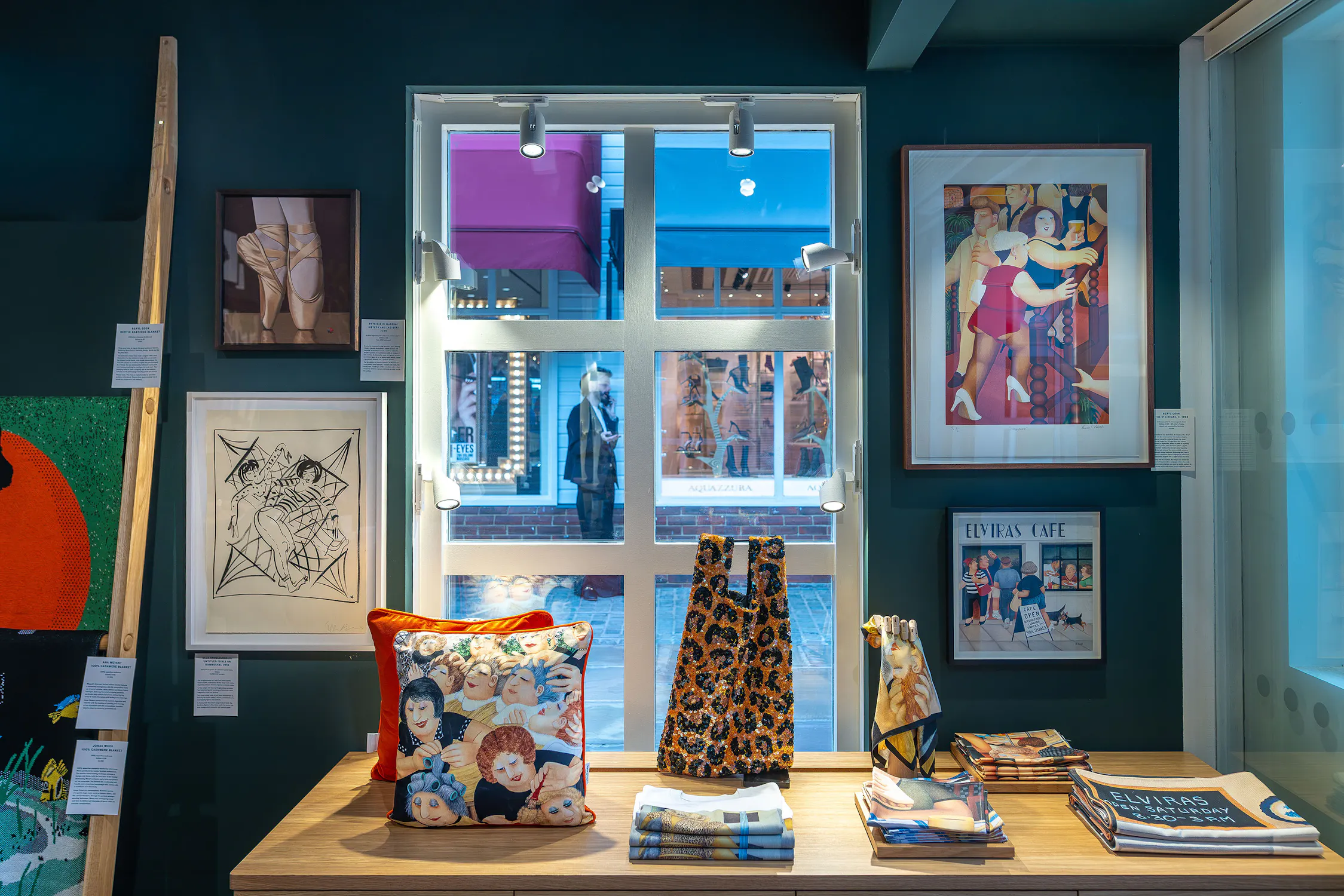 The interior of the House of Voltaire pop-up at Bicester Village displays framed art, colourful cushions, and folded textiles on a wooden table. A large window overlooks a busy street. Artworks feature abstract and figurative designs. The scene is bright and cosy, with modern decor and various patterns on textiles.