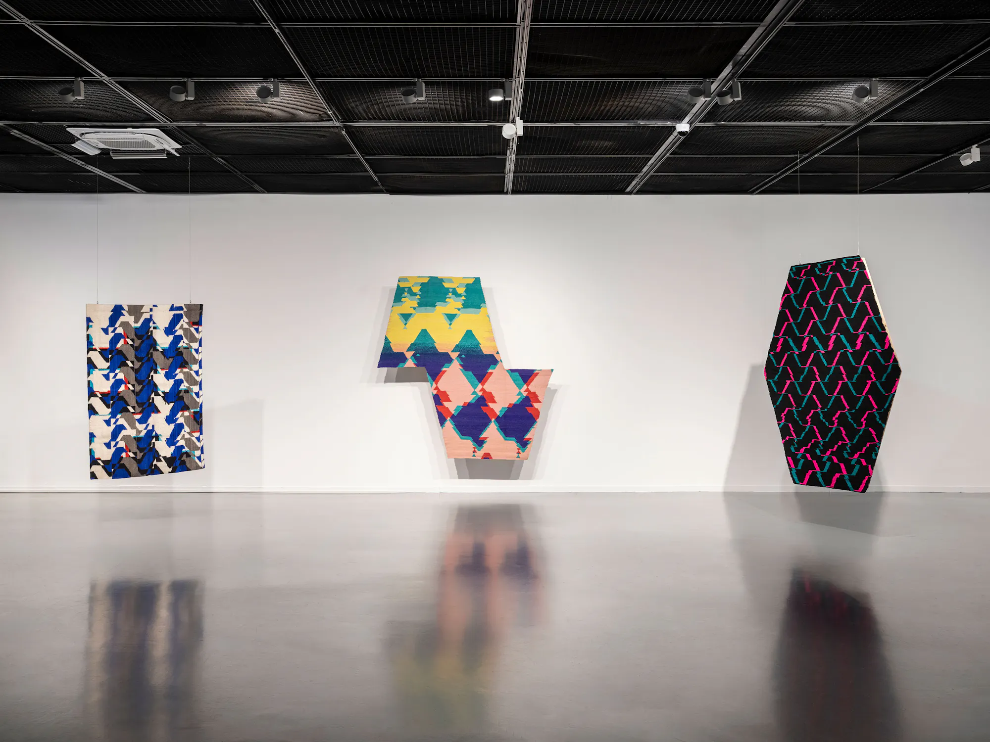 Three abstract, geometric textile pieces by GUido Yannitto with zigzag patterns in blue, green, yellow, red, pink, and black hang on a white gallery wall. The artworks are irregular in shape and cast soft shadows on the clean, polished floor.