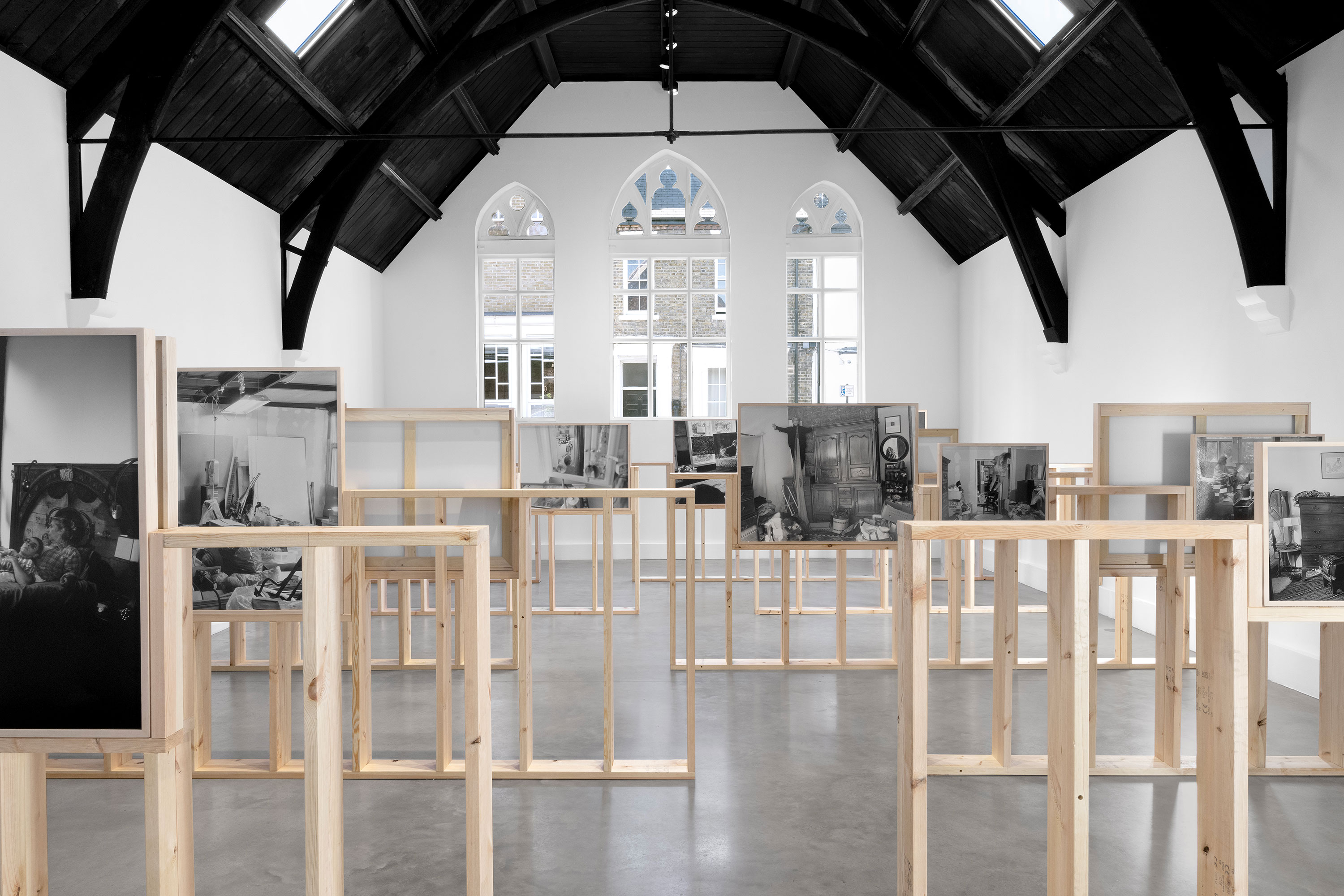 An art gallery features multiple framed black-and-white photographs by Katherine Hubbard mounted on freestanding wooden frames. The room has high vaulted ceilings with dark wooden beams and large arched windows at the far end.