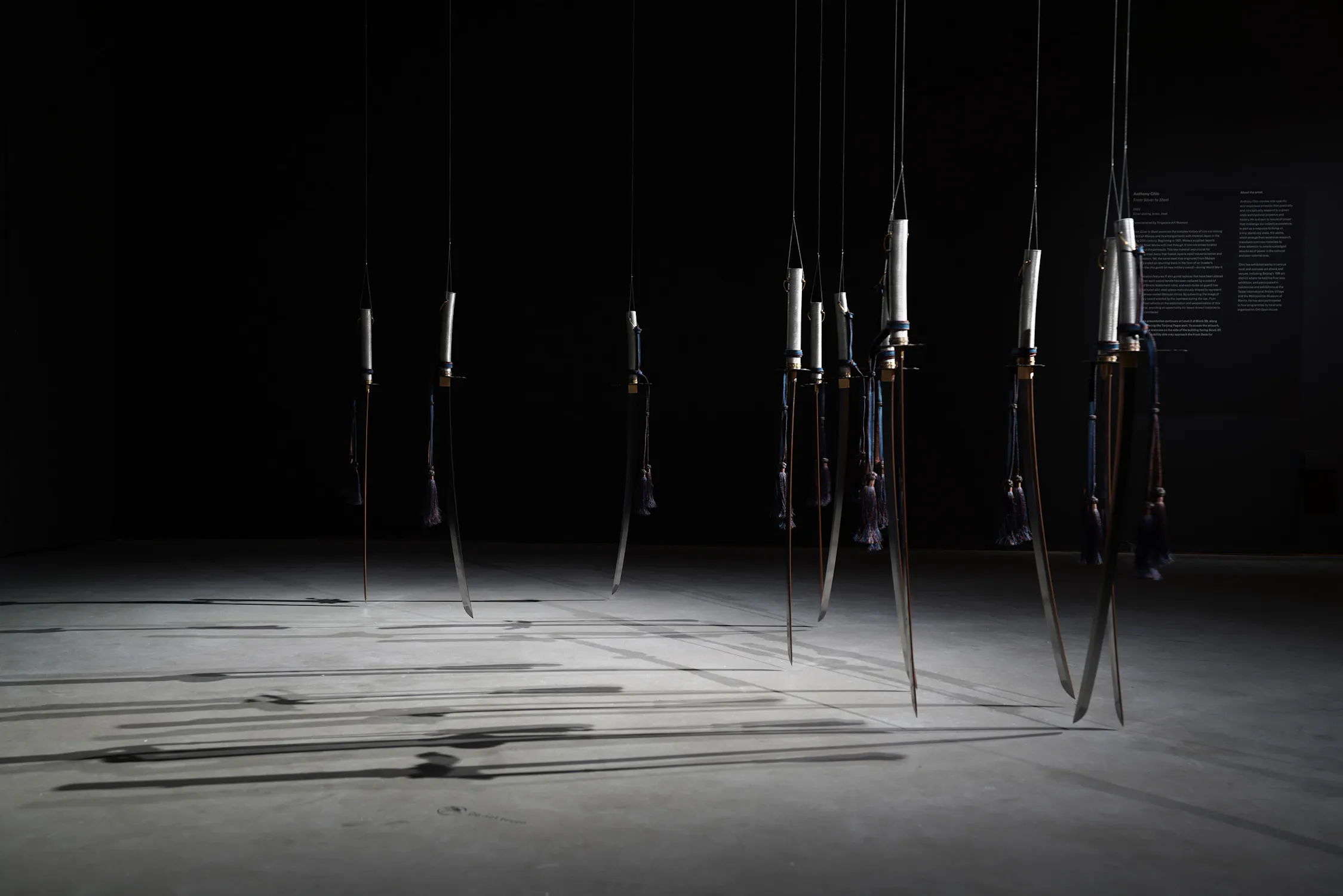 Several Japanese swords hang vertically from the ceiling, their blades pointing towards a concrete floor. Each handle features a decorative tassel, casting long shadows across the otherwise dark, gallery-like space.