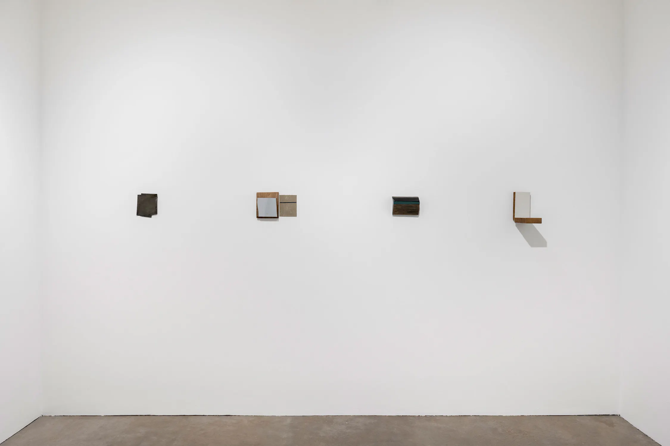Four small, abstract minimalist sculptures by Bill Walton are mounted in a horizontal row on a large, white gallery wall. Each piece features simple geometric forms in muted tones of brown, grey, and black, casting soft shadows against the plain background.