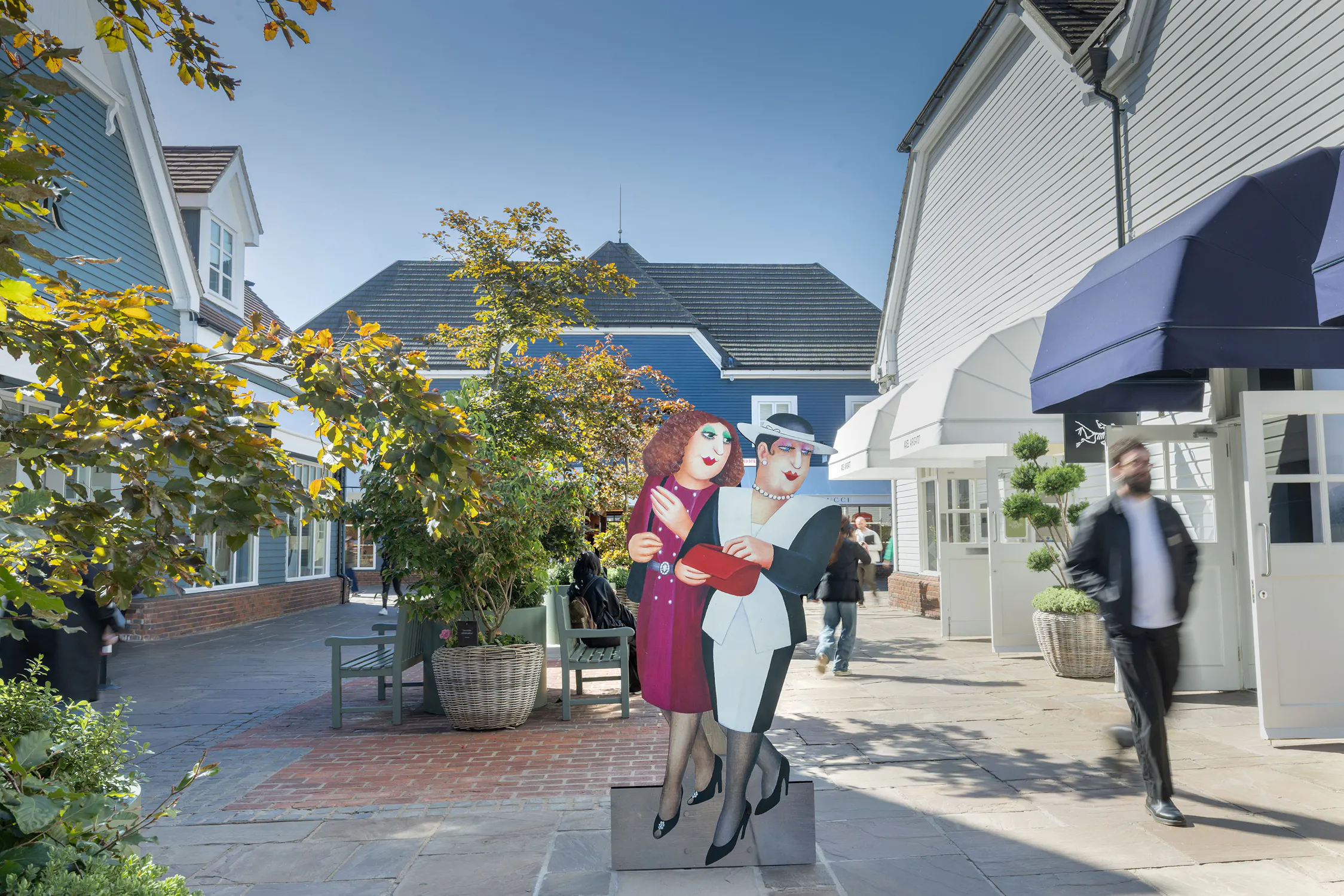 A colourful cut-out of two glamourously dressed women stands in a sunny outdoor shopping area with blue-and-white shopfronts, green plants, and people walking by. The sky is clear and blue, and there are shadows on the ground.