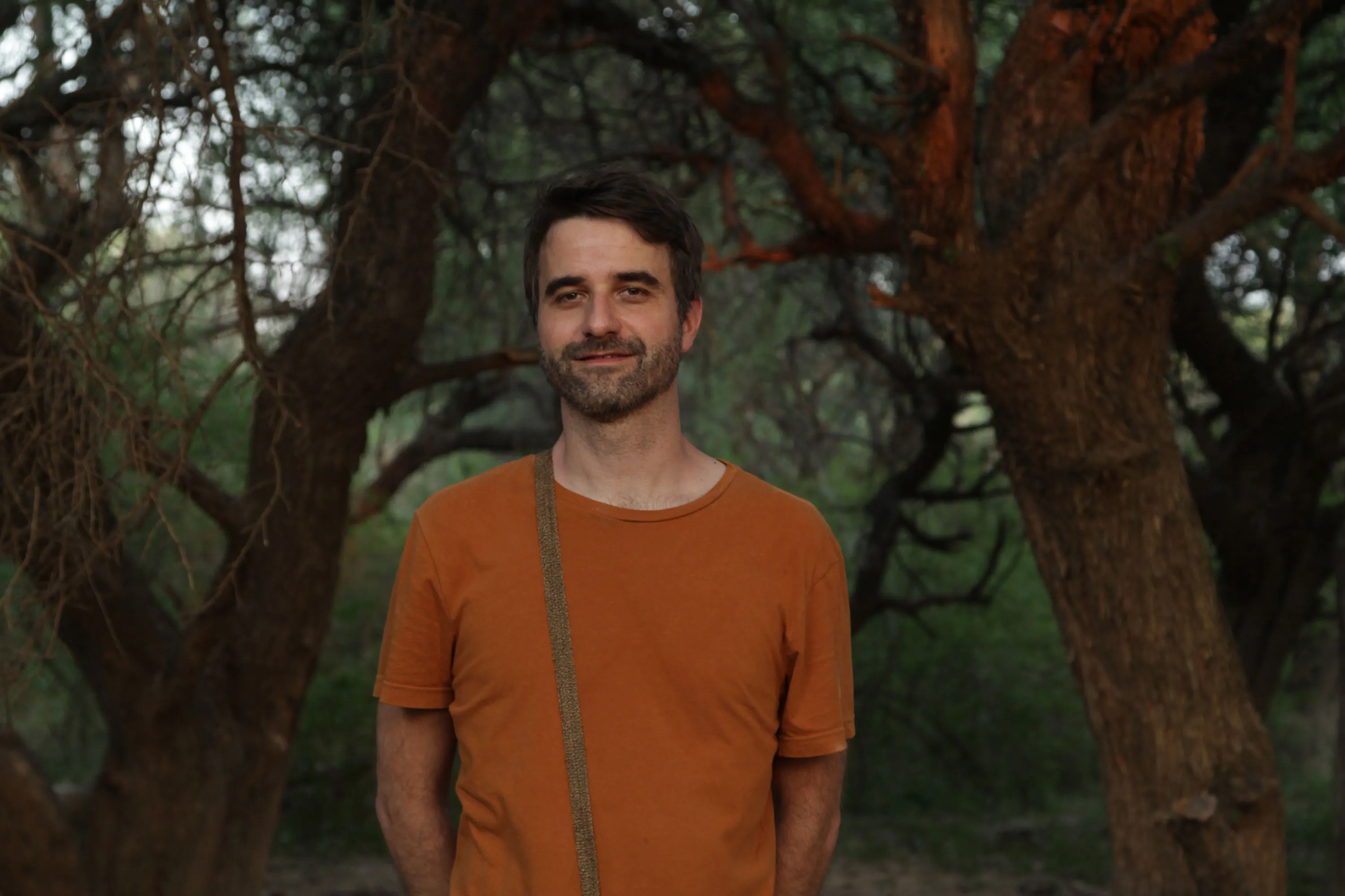 Portrait of artist Guido Yannitto. A man with short dark hair and a beard stands outdoors among trees, wearing an orange T-shirt and a crossbody strap. He is smiling slightly, with his hands behind his back. The background is lush and green, with soft natural light.