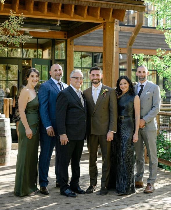 Outdoor family portrait with relatives in suits and dresses in front of a rustic home.