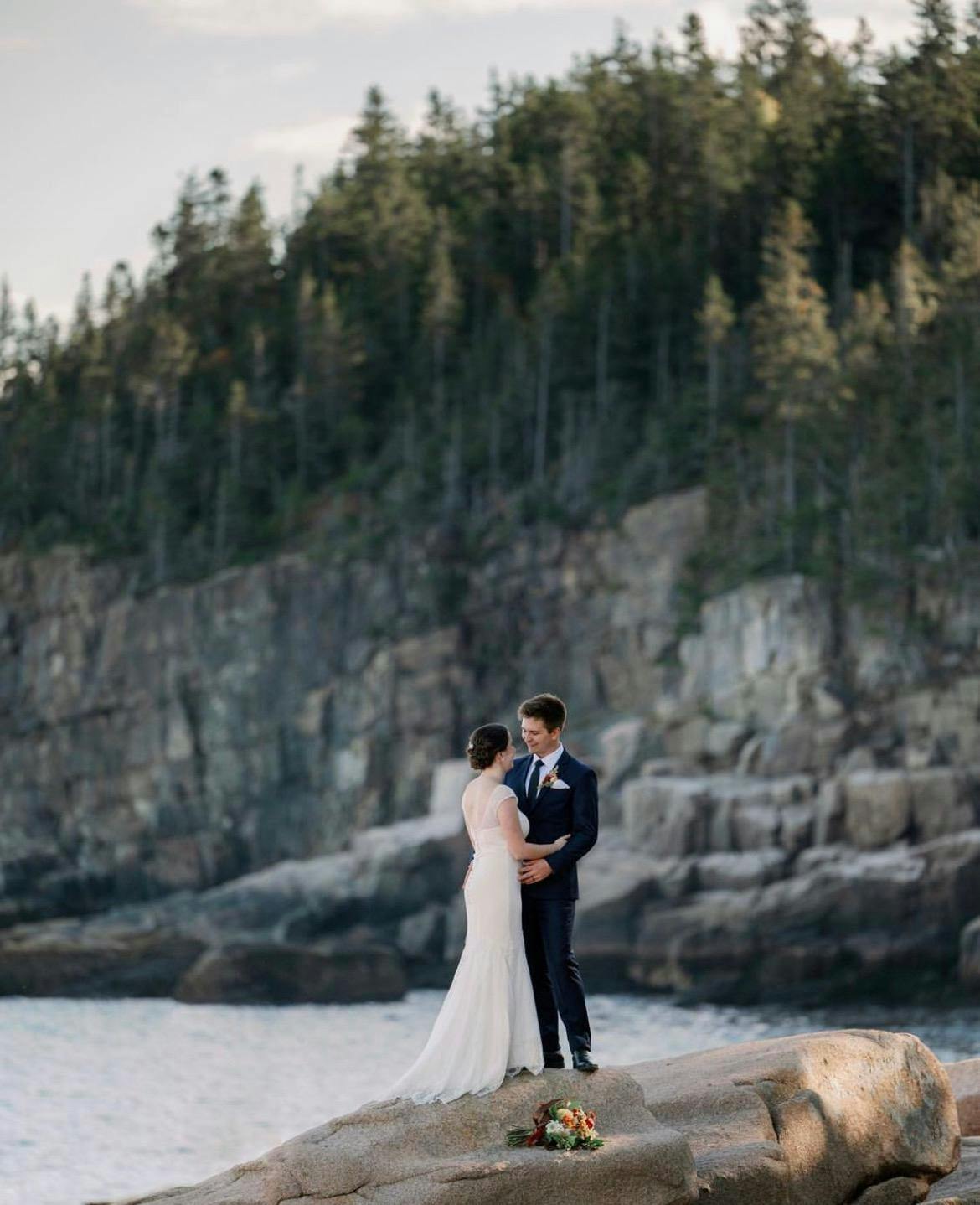A newlywed couple poses outdoors beside a calm lakeside, surrounded by natural greenery and soft light, capturing a romantic outdoor wedding moment.