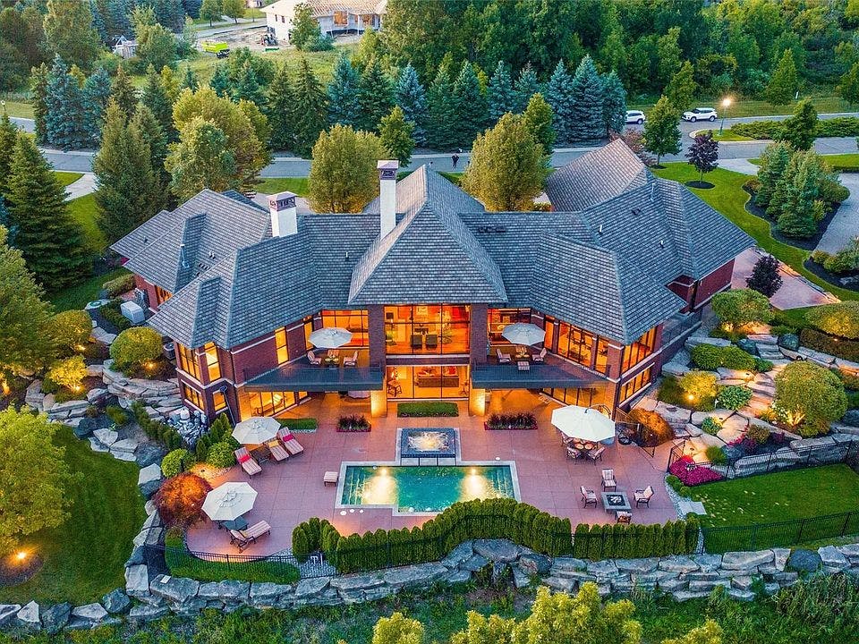A spacious Oakland Township, Michigan mansion with large windows, a landscaped yard, a sparkling pool, sun beds, and surrounding green pine trees.