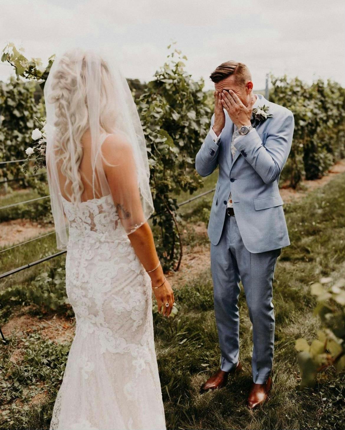 A groom gently covering his eyes with his hands as his bride stands before him in a lush garden filled with greenery, white roses, and soft grass, capturing an intimate wedding moment.