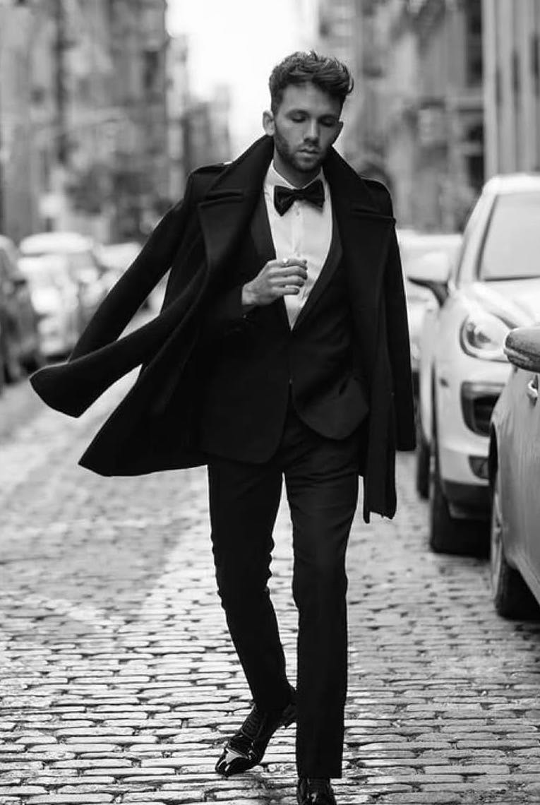 A stylish man in a black tuxedo and bow tie walks confidently down a cobblestone city street, wearing a flowing overcoat in a black-and-white fashion editorial setting.