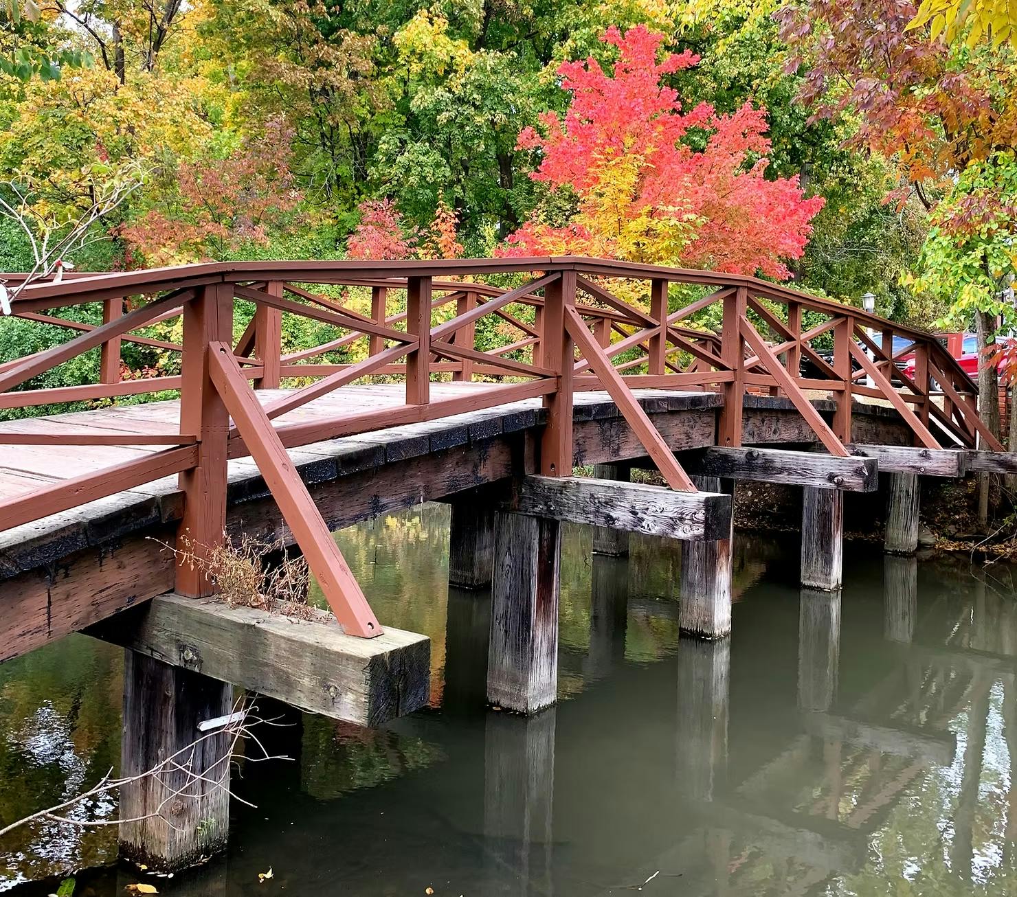 Scenic view of a wooden bridge in Northville, Michigan during fall, with colorful autumn leaves on trees and reflected in the calm water beneath the bridge.