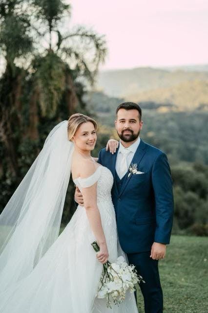 Bride and groom posing outdoors, the groom wearing a blue suit with a white tie, surrounded by greenery and soft natural light, capturing a romantic wedding moment.