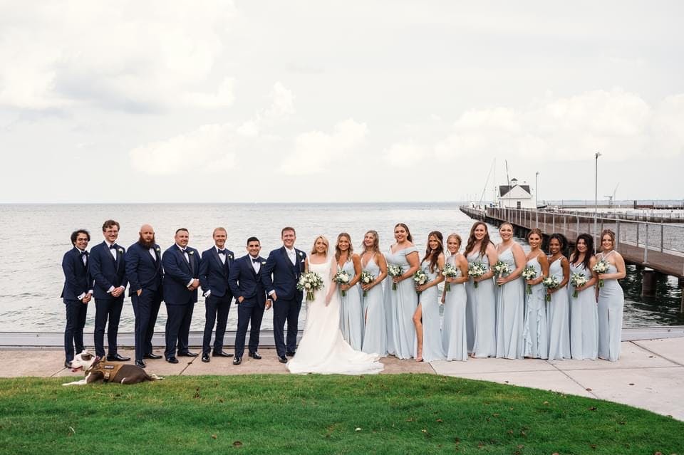 Bridesmaids and groomsmen posing outdoors in front of a serene lake, dressed elegantly for the wedding, capturing a joyful group moment with natural scenic beauty in the background.
