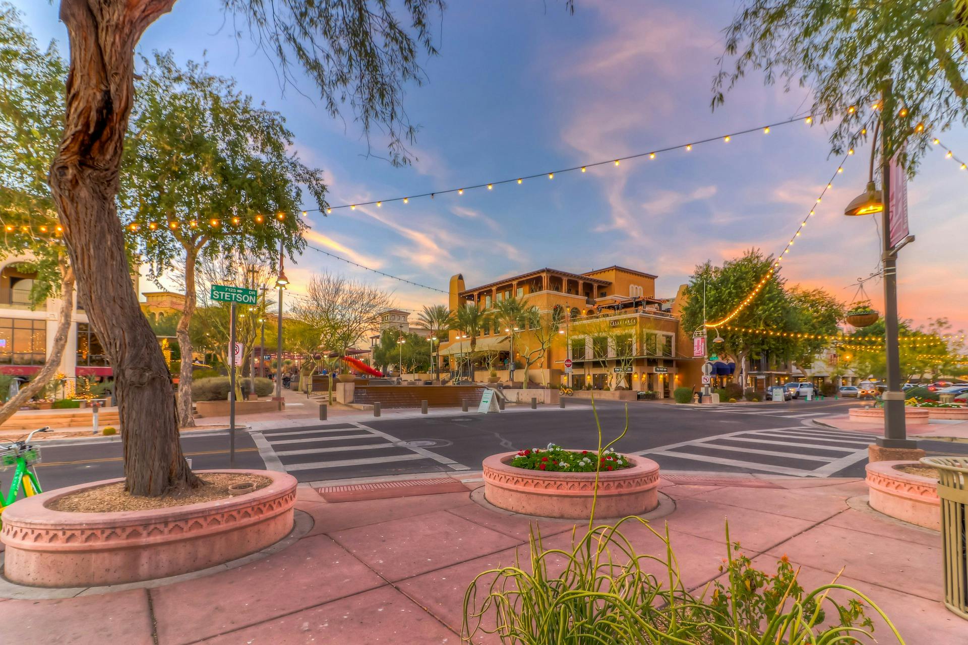 Vibrant downtown street scene at sunset with warm desert-style buildings, palm trees, and string lights glowing above the sidewalks near Stetson Drive, capturing a lively and colorful urban atmosphere.