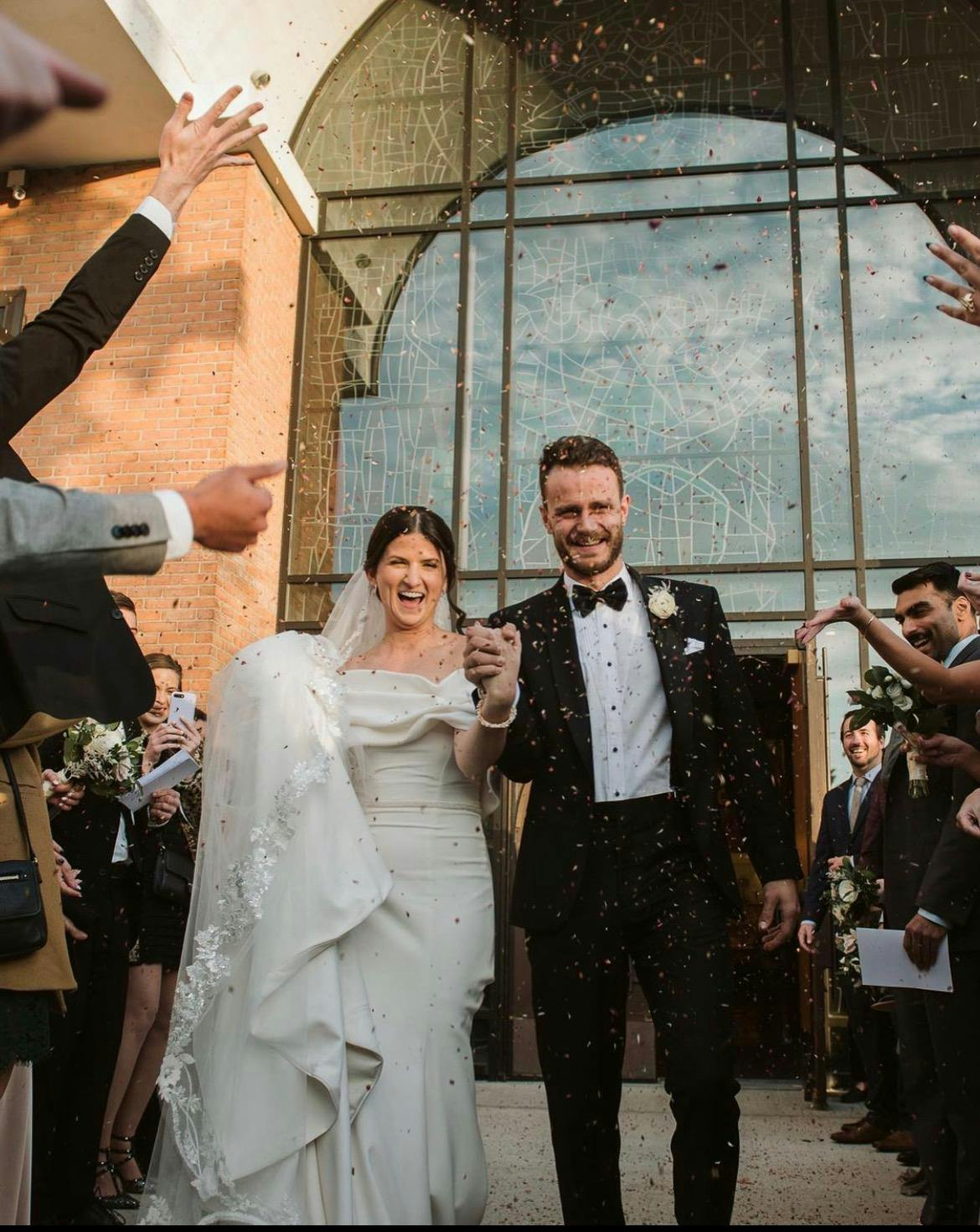 Joyfully tossing confetti over bride and groom dressed in a classic tuxedo, capturing a fun and celebratory wedding moment filled with movement and laughter.