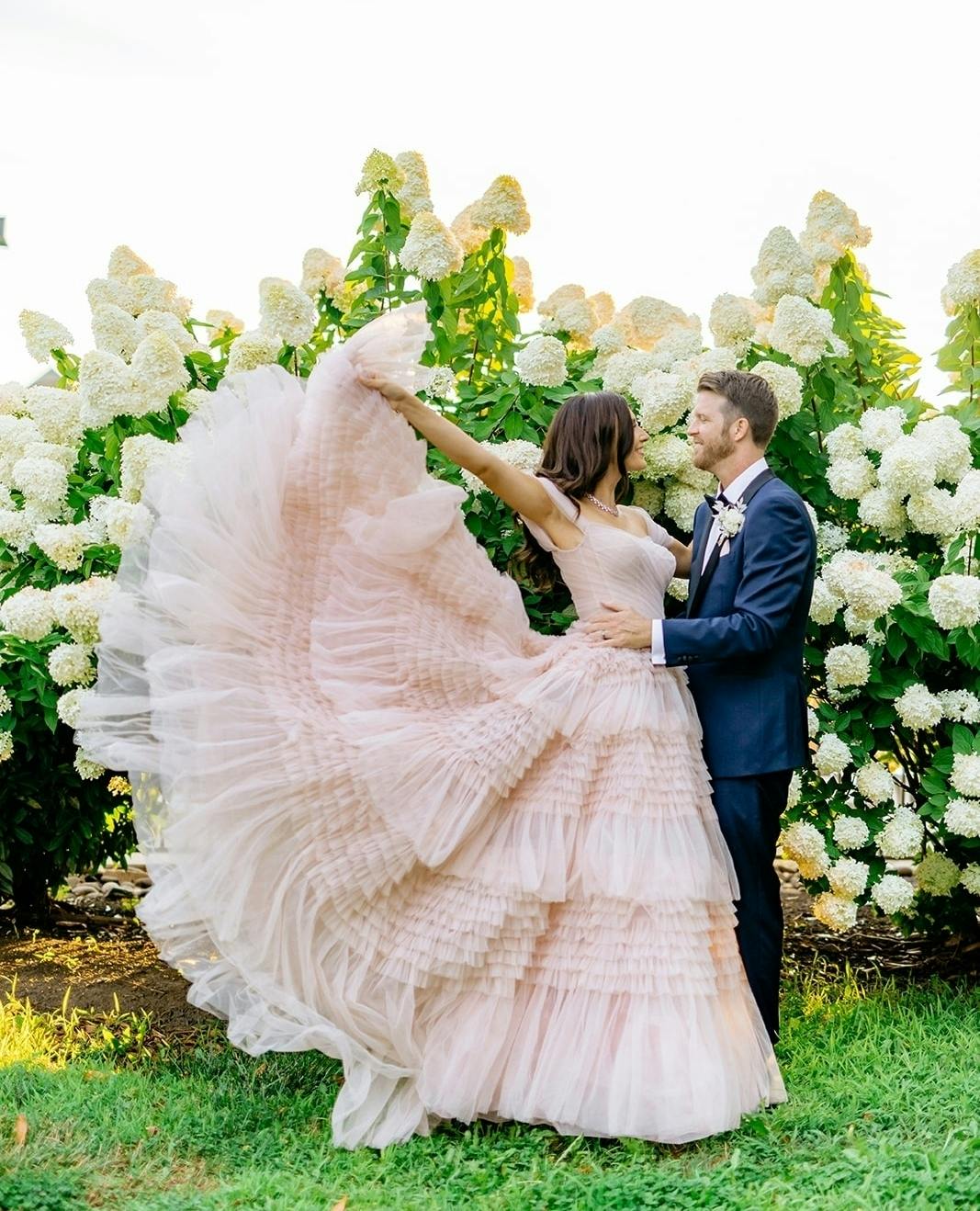 Bride wearing blush wedding gown with layered tulle ruffle layers and ruched bodice and groom wearing navy tuxedo for garden wedding.