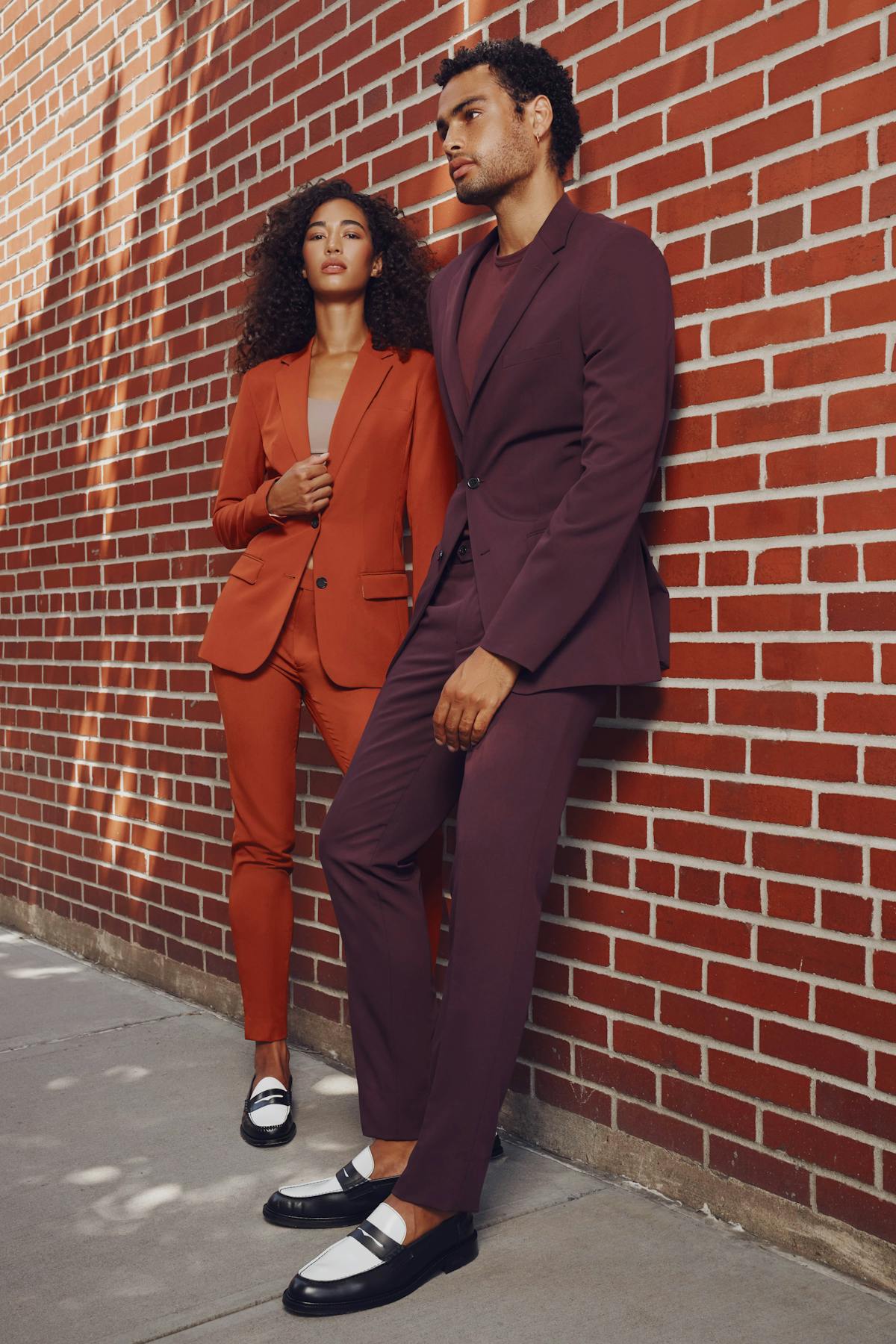 Monochrome suit outfits with simple styling in a women's burnt orange suit and a men's burgundy suit against a brick wall.