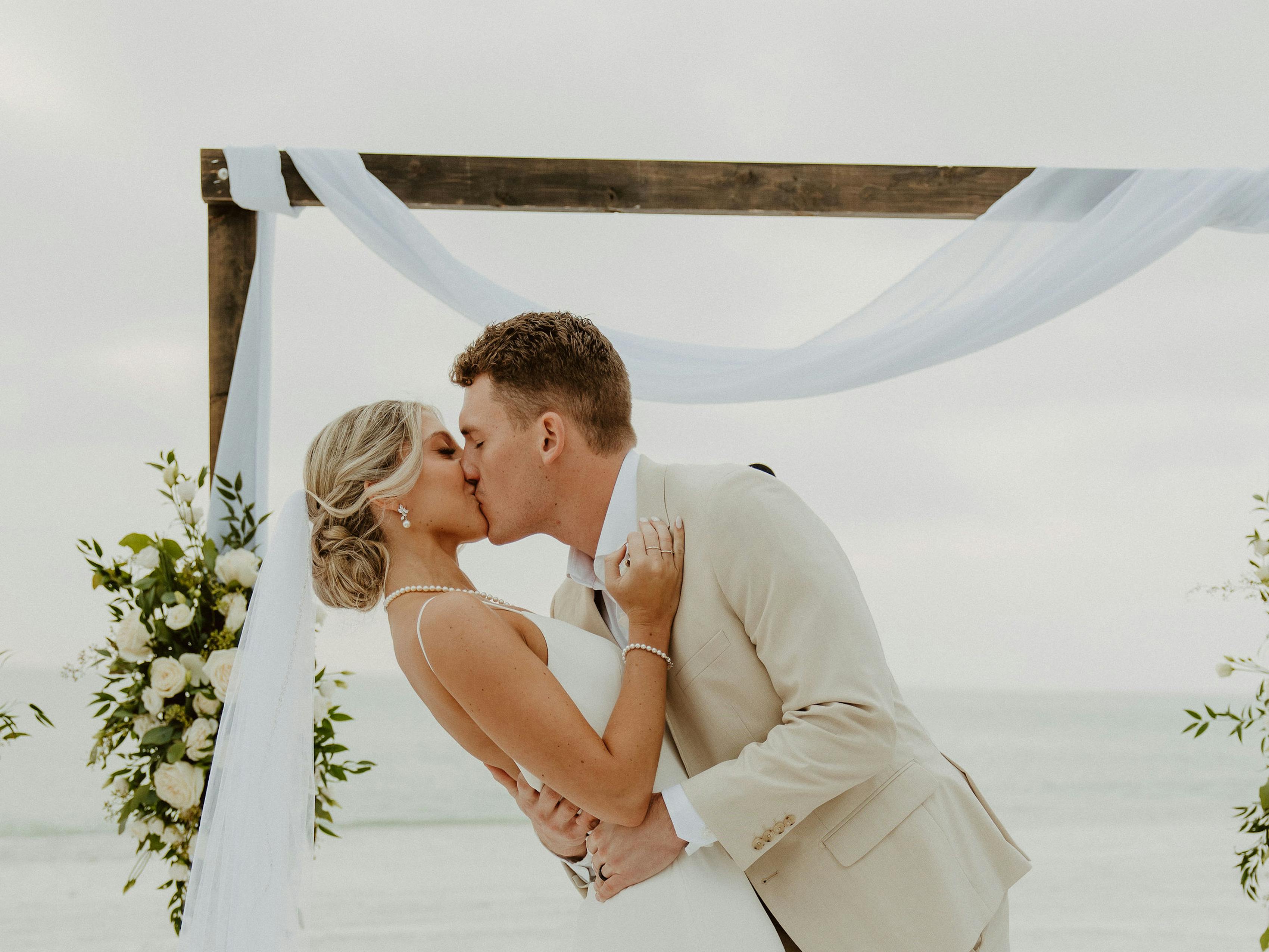 Bride in simple white dress and groom in tan suit at destination beach wedding kissing at the altar.