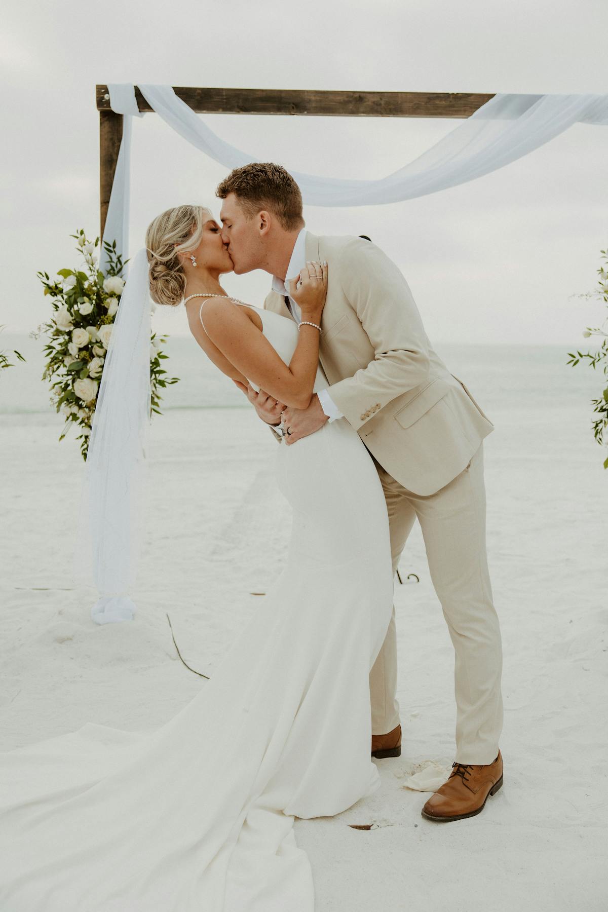 Bride in simple white dress and groom in tan suit at destination beach wedding kissing at the altar.