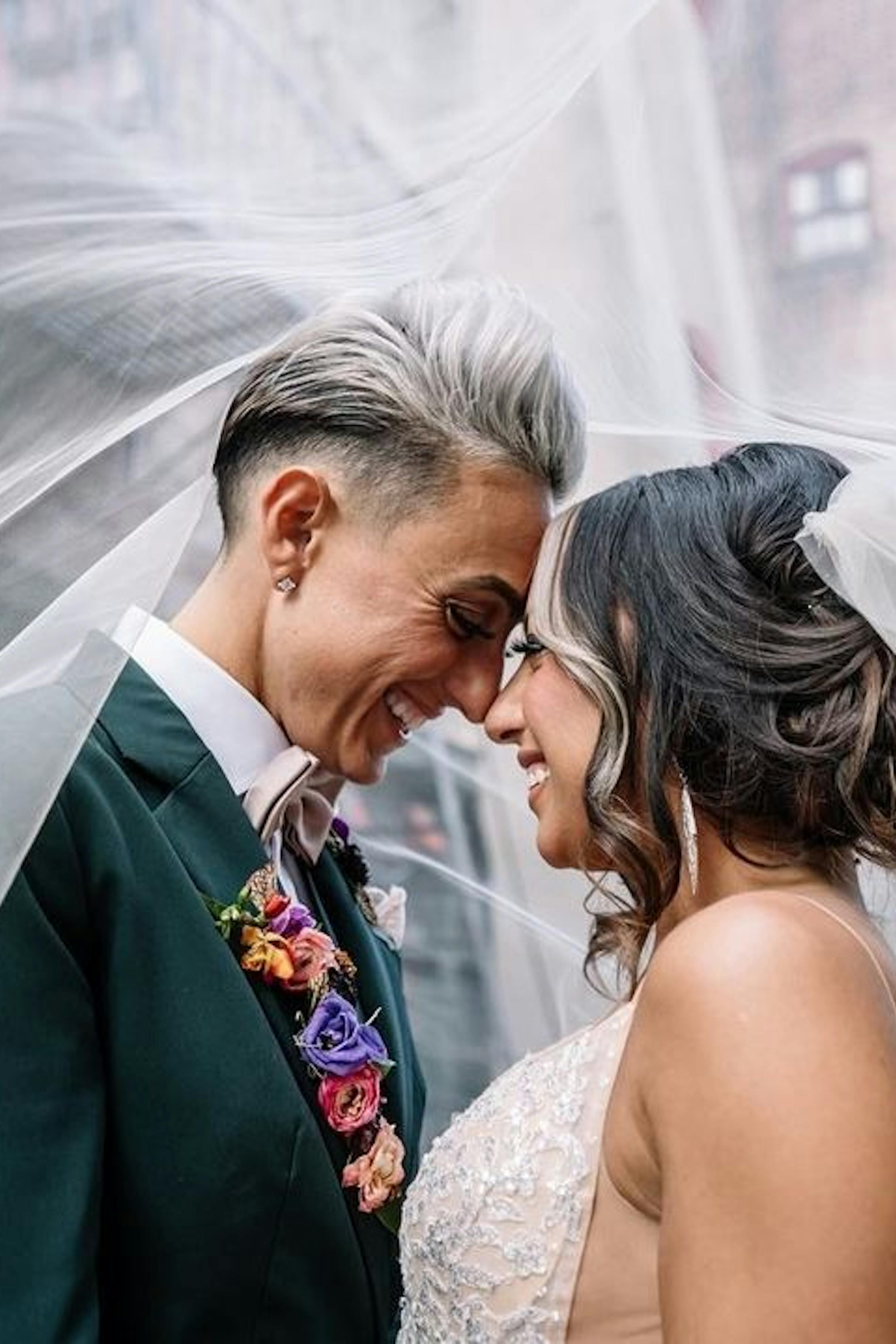 Lesbian brides on their wedding day surrounded by wedding veil; one bride wears dark green women's suit with rainbow lapel flowers and blush bow tie.