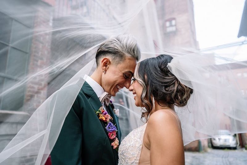 Lesbian brides on their wedding day surrounded by wedding veil; one bride wears dark green women's suit with rainbow lapel flowers and blush bow tie.