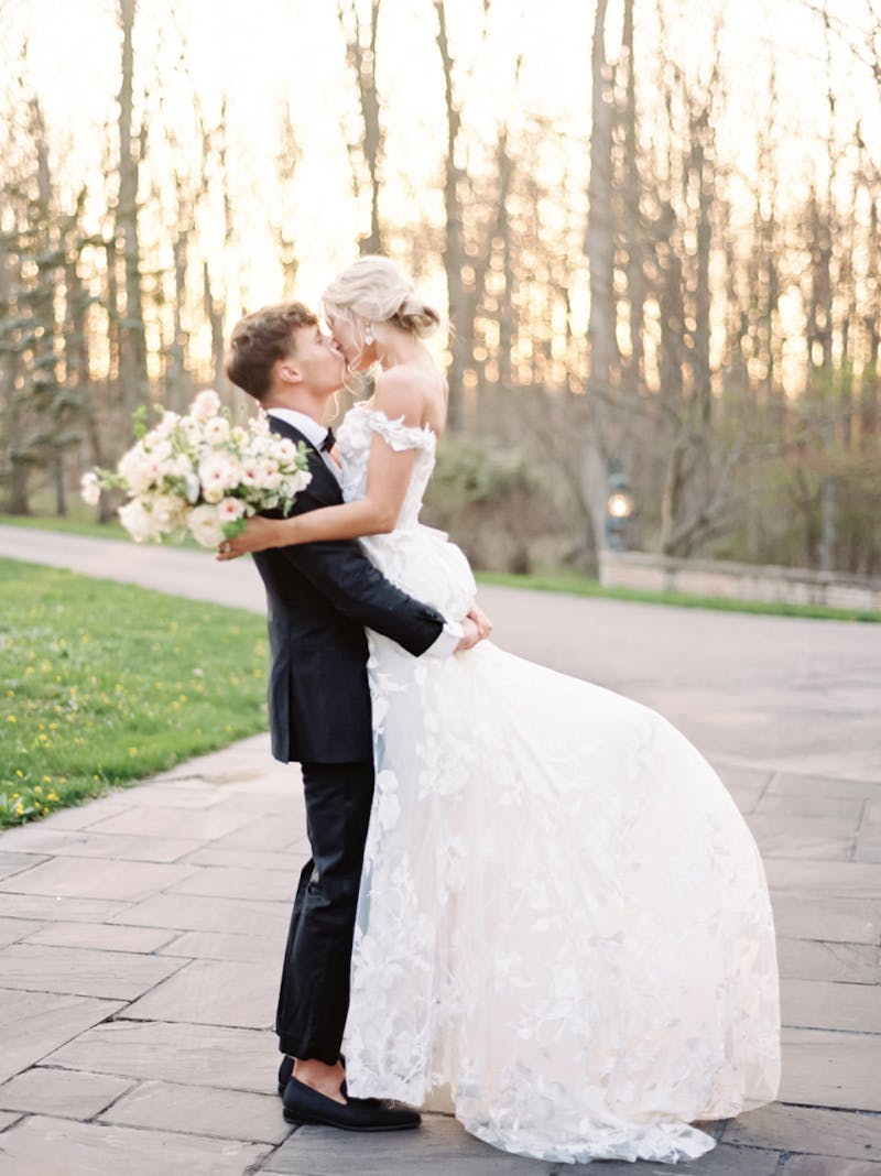 Romantic European theme groom wearing classic slim fit black tuxedo at wedding lifting bride in white lace ballgown.