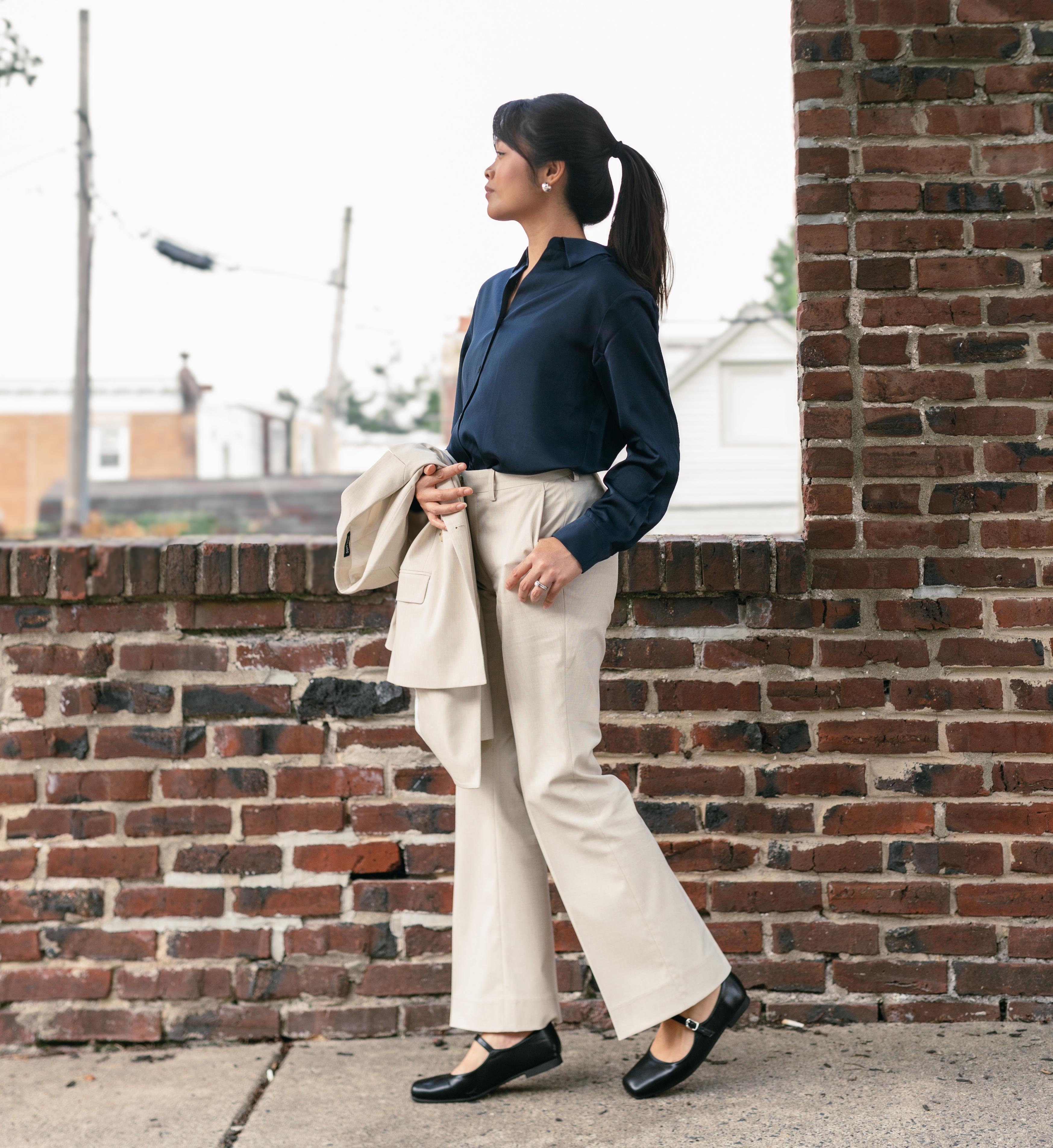 Woman wearing a navy blouse with cream wide-leg trousers and black flats, holding a matching cream blazer for a casual wedding outfit.