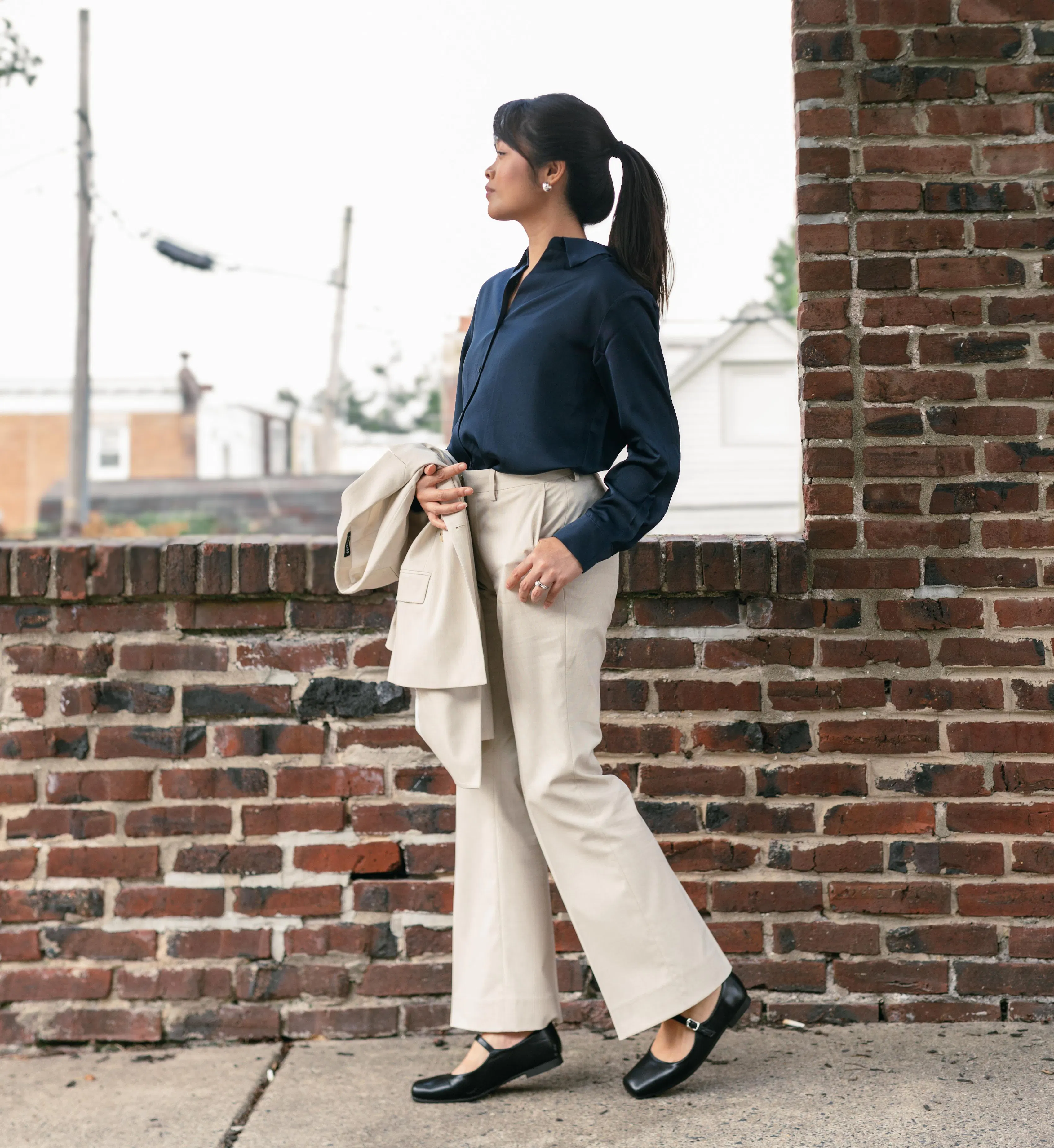 Woman wearing a navy blouse with cream wide-leg trousers and black flats, holding a matching cream blazer for a casual wedding outfit.