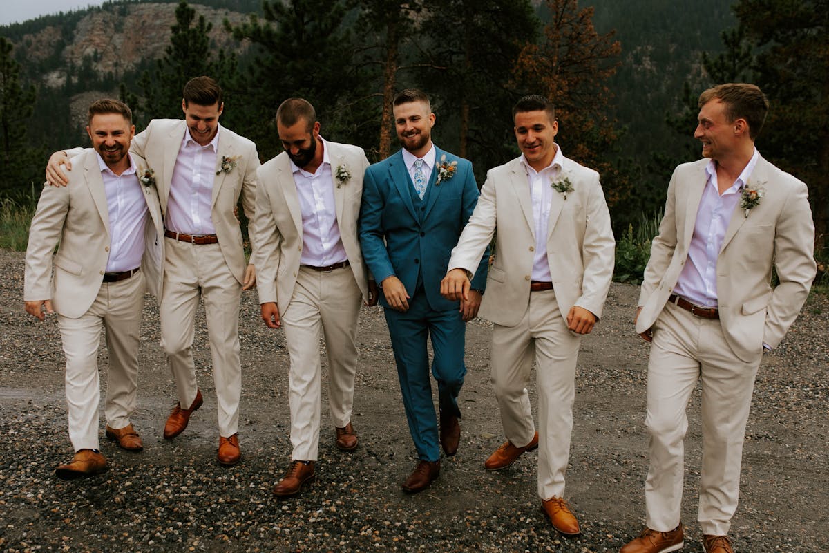 Groom standing out from groomsmen wearing a 3 piece blue suit and suit vest surrounded by light tan groomsman suits with no tie.
