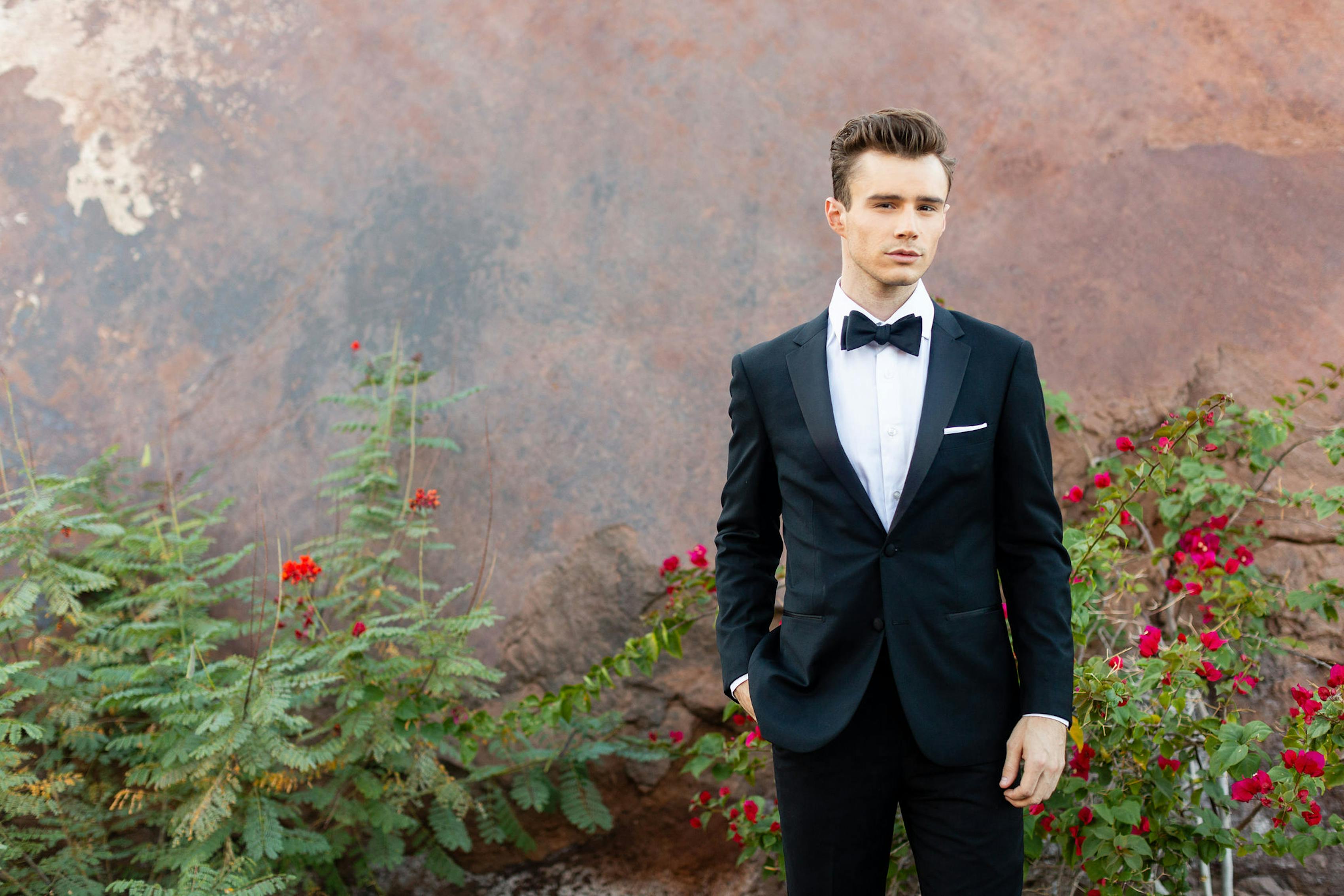Man in black tie wedding guest wearing outfit wearing a notch lapel black tuxedo with traditional tuxedo accessories at formal outdoor event with roses.