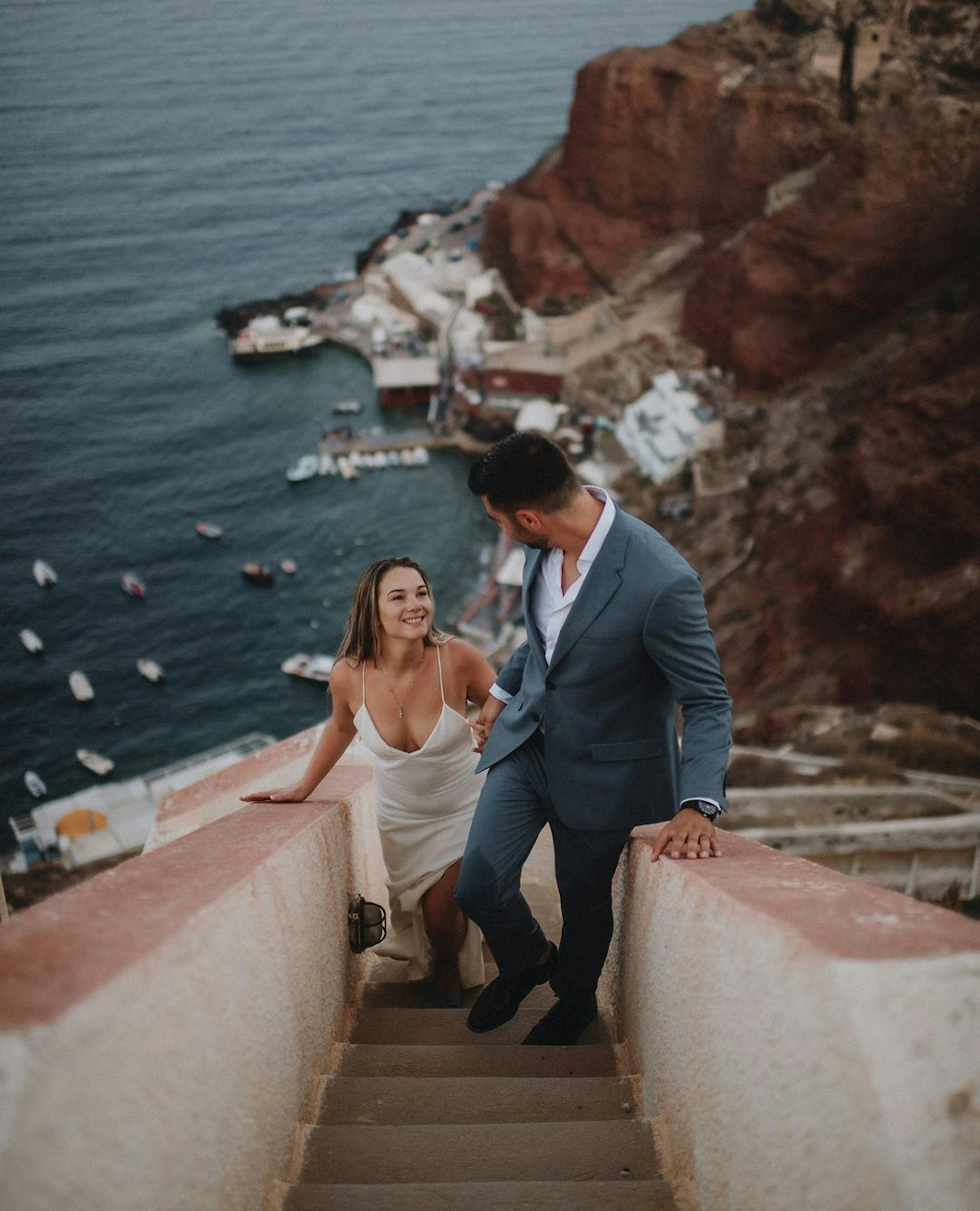 Bride wearing slinky simple lightweight wedding dress holding hands with groom wearing light blue beachy wedding suit walking up stairs in Greece.