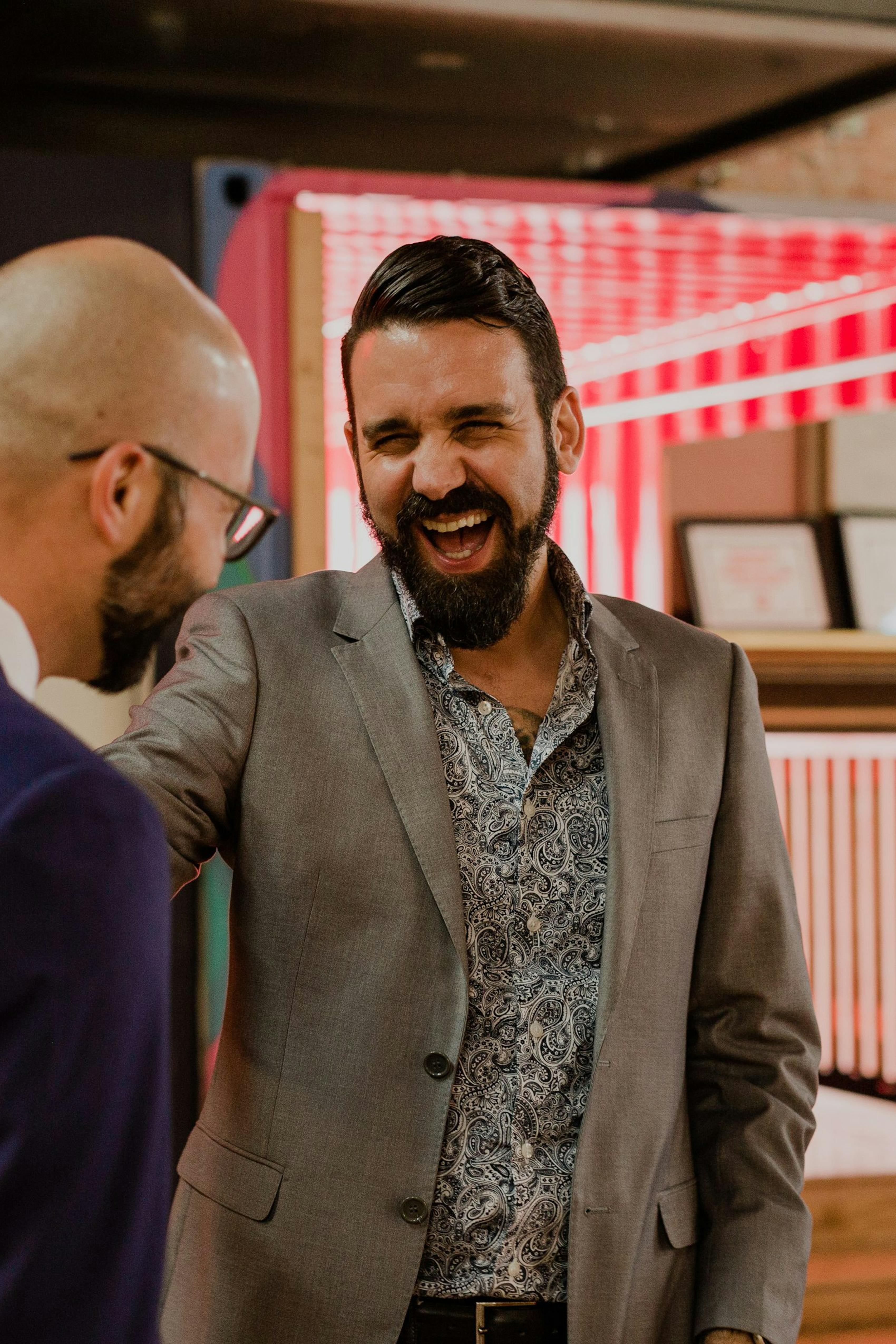 Two men wearing suits laughing, one in a light grey suit with paisley patterned dress shirt and one in a navy blue men's suit jacket.