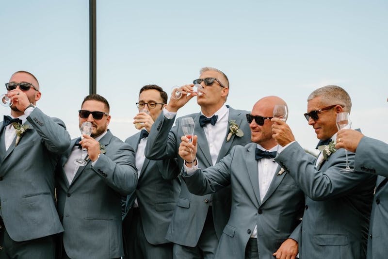 Group of groomsmen wearing light grey wedding suits with sunglasses and navy bowties holding champagne to cheers.
