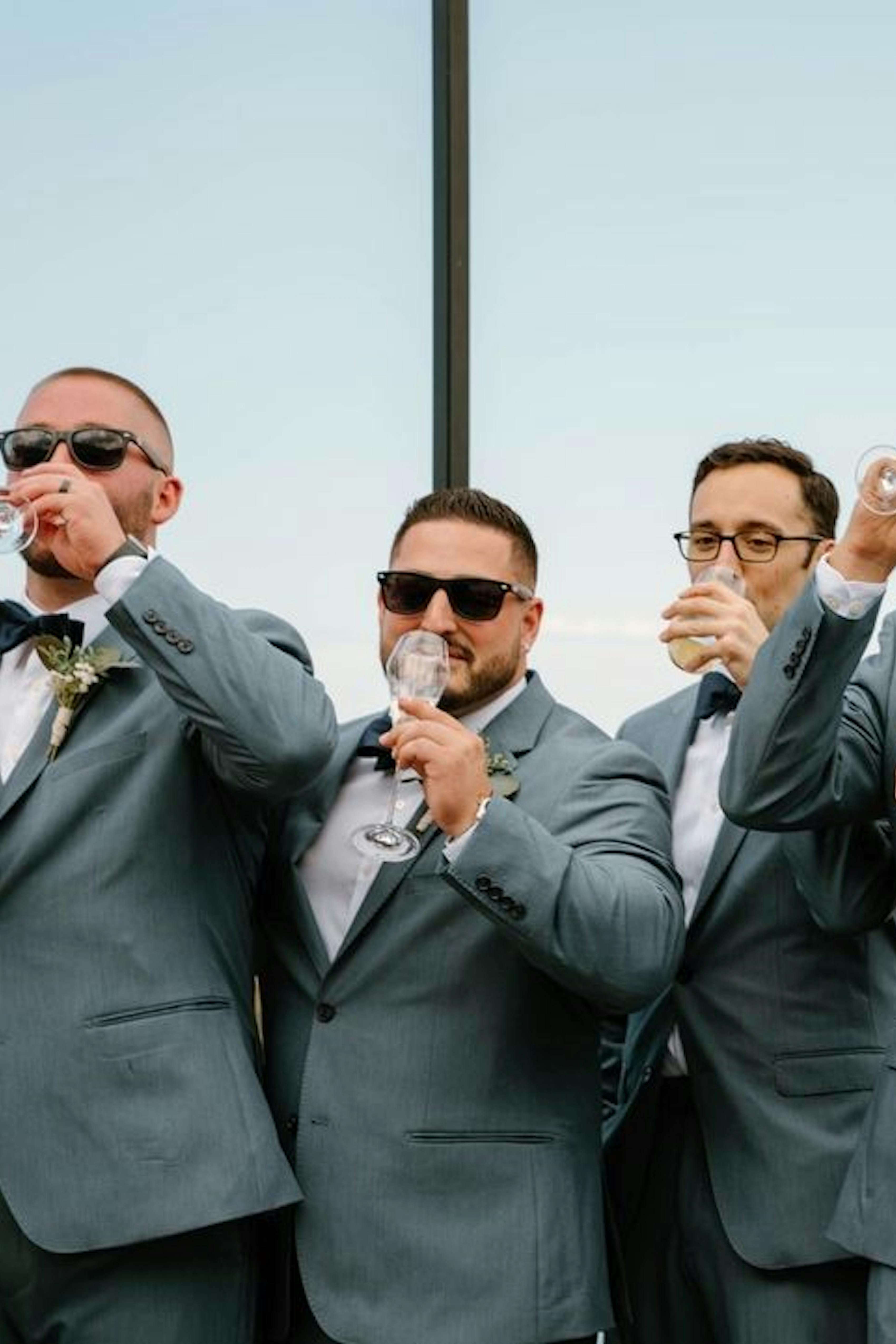 Group of groomsmen wearing light grey wedding suits with sunglasses and navy bowties holding champagne to cheers.