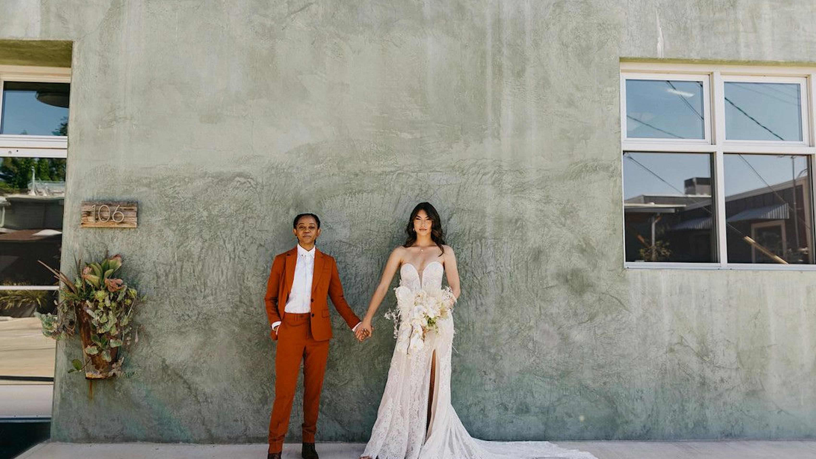 Wedding dress alternative for the edgy bride women's burnt orange suit alongside bride in alternative off-white wedding dress at industrial-chic boho wedding venue.