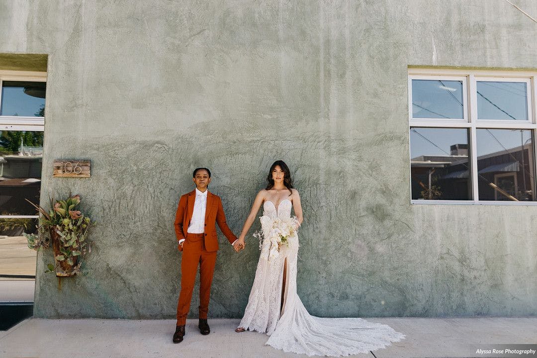 Wedding dress alternative for the edgy bride women's burnt orange suit alongside bride in alternative off-white wedding dress at industrial-chic boho wedding venue.