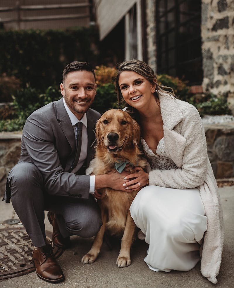 Winter wedding attire with groom wearing light grey wedding suit and bride wearing lace wedding dress and boucle overcoat hugging golden retriever at wedding.