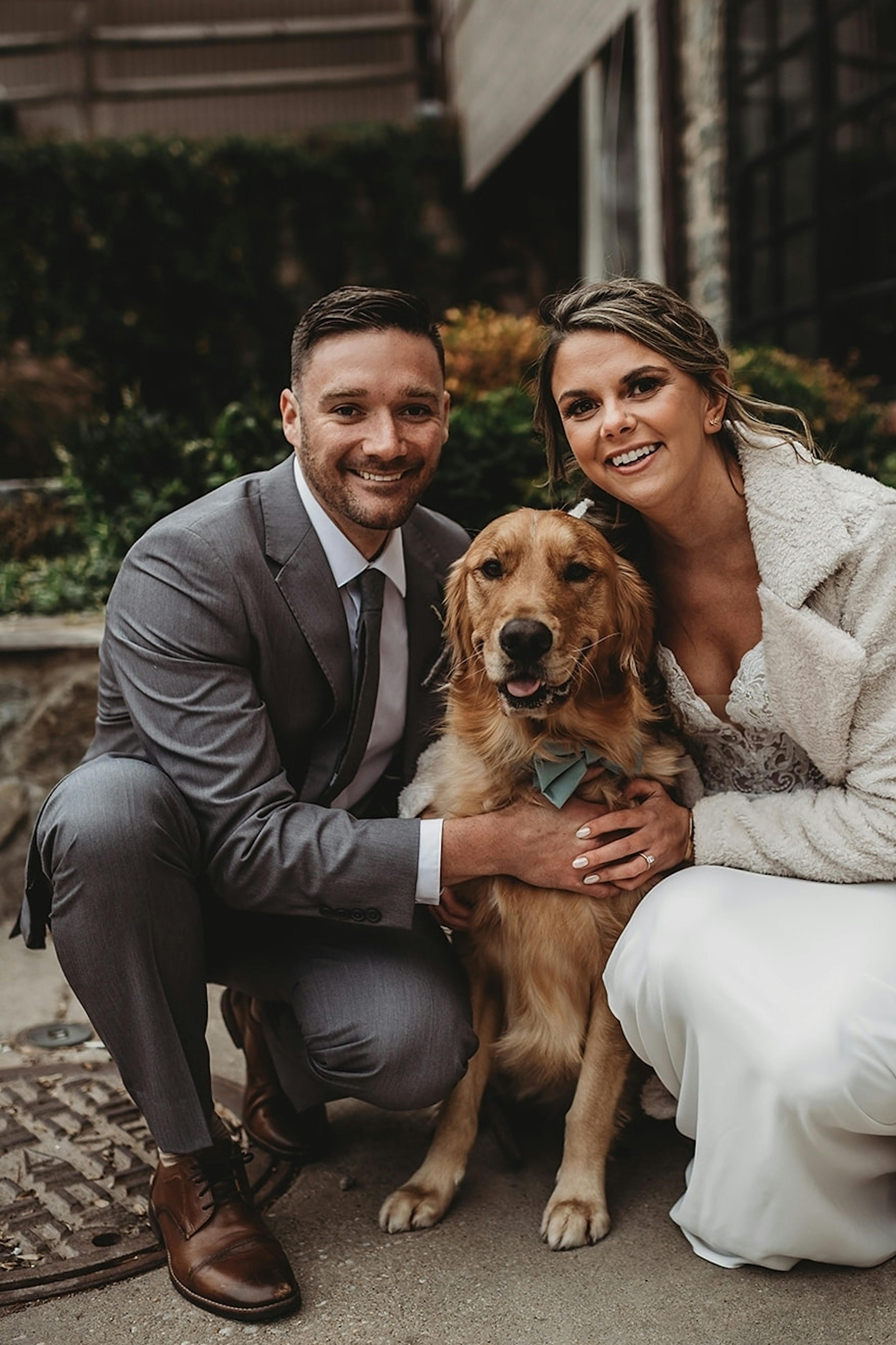 Winter wedding attire with groom wearing light grey wedding suit and bride wearing lace wedding dress and boucle overcoat hugging golden retriever at wedding.