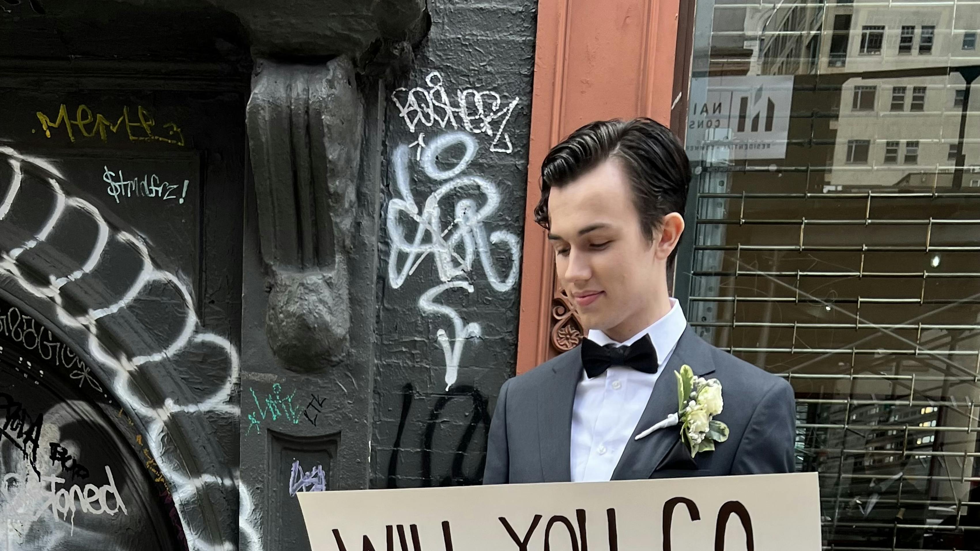 High school promposal sign idea dressed up wearing gray prom suit on city street.