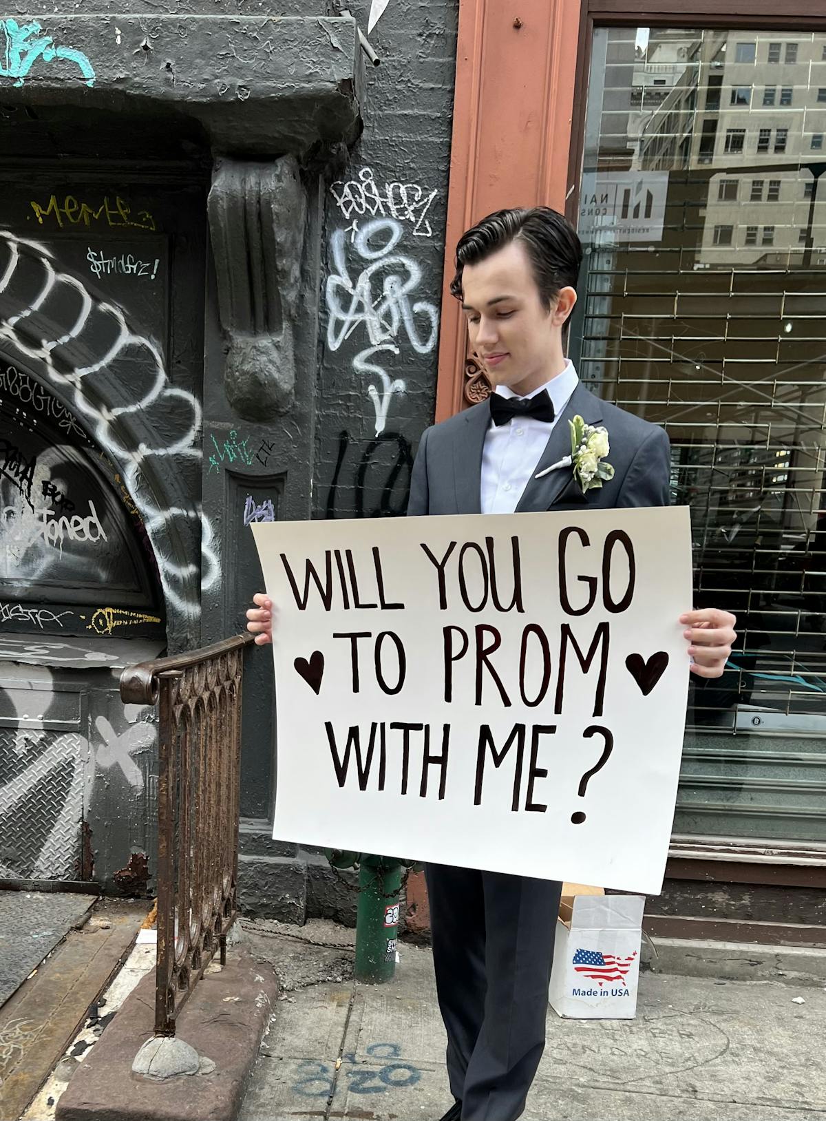 High school promposal sign idea dressed up wearing gray prom suit on city street.